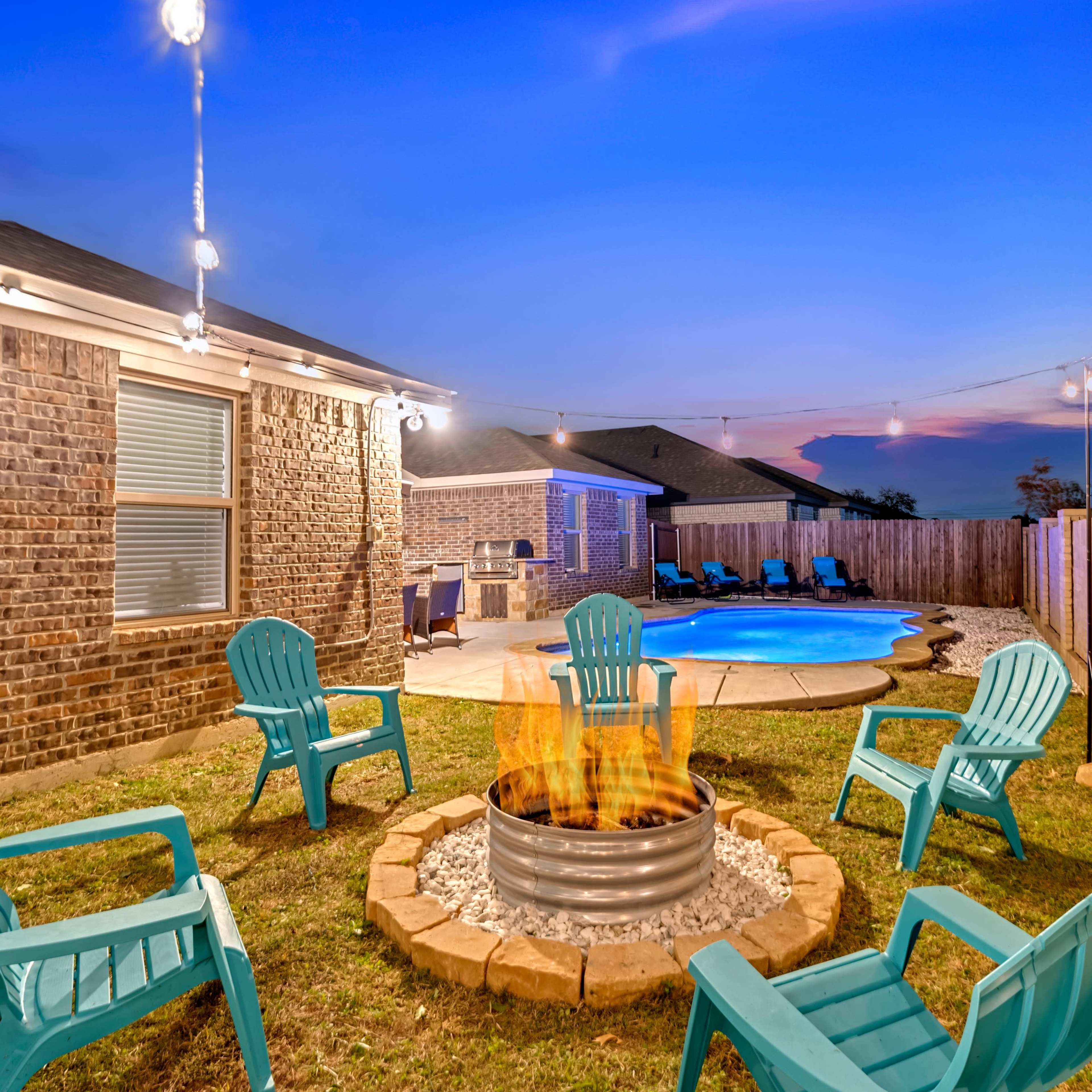 A fire pit surrounded by blue Adirondack chairs sits in a backyard with a pool and string lights strung overhead at dusk.
