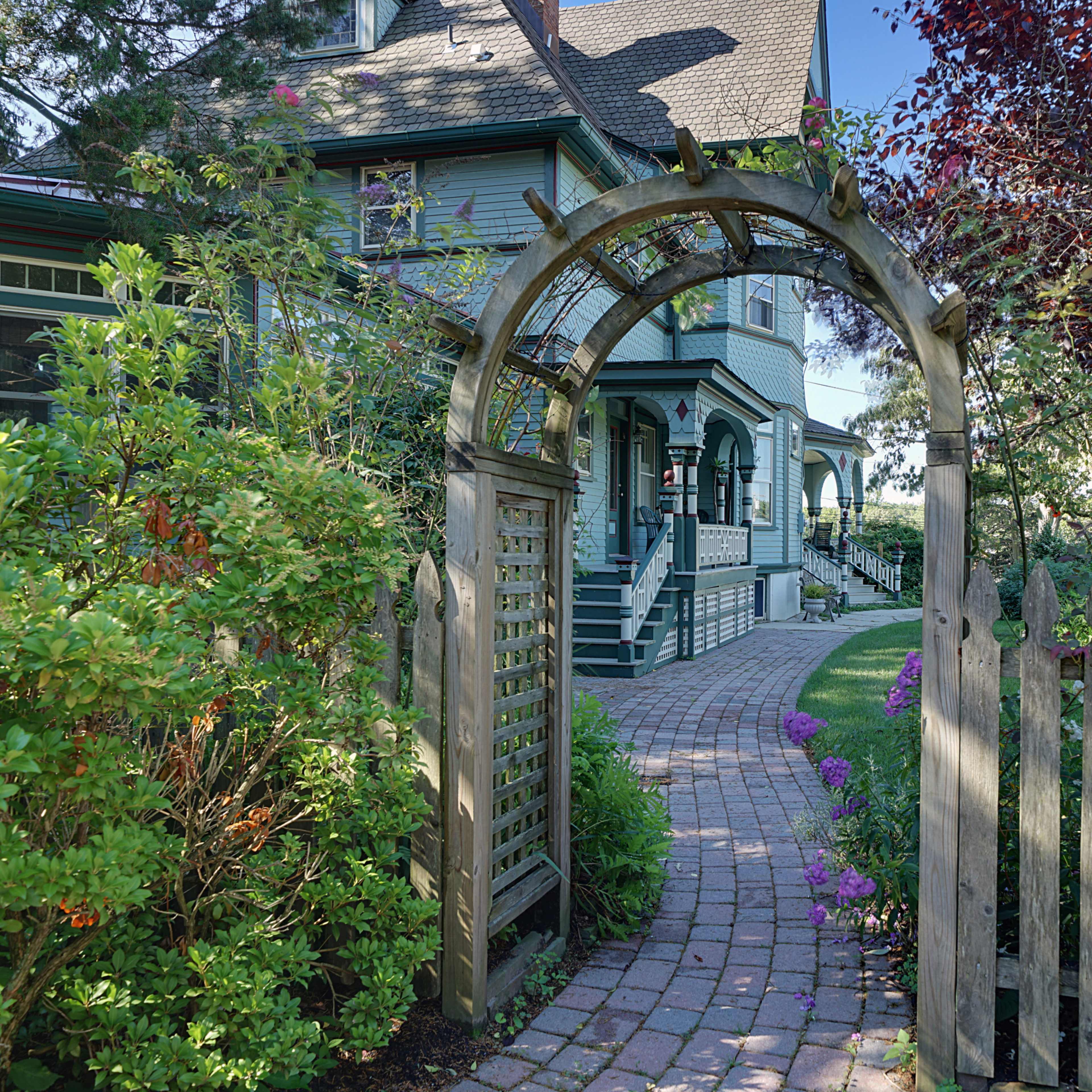 A wooden archway leads to a path of brick pavers bordered by flowers, leading to a house with a green exterior and surrounding gardens.