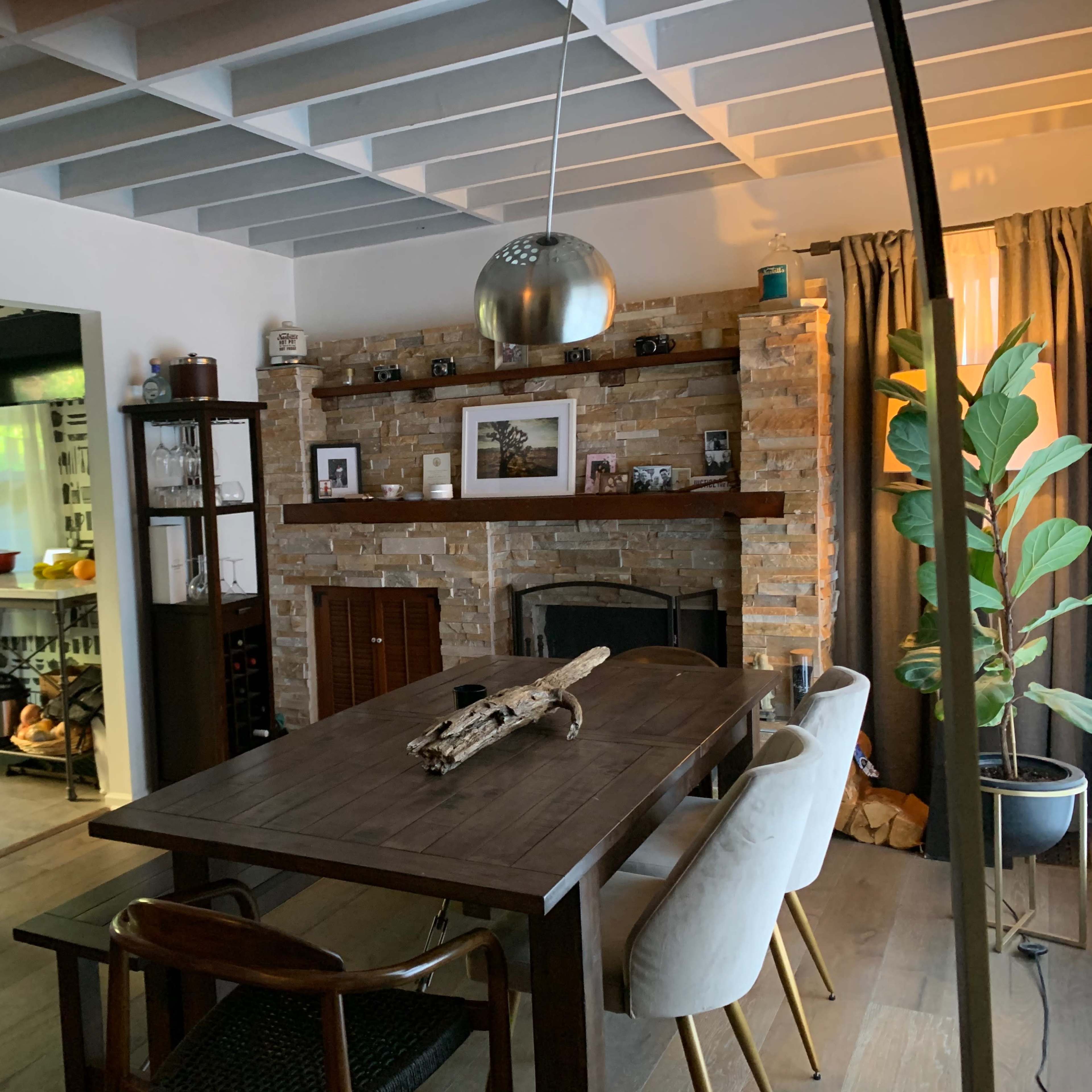 A dining area with a wooden table, surrounded by chairs, next to a stone fireplace, and an open kitchen in the background.
