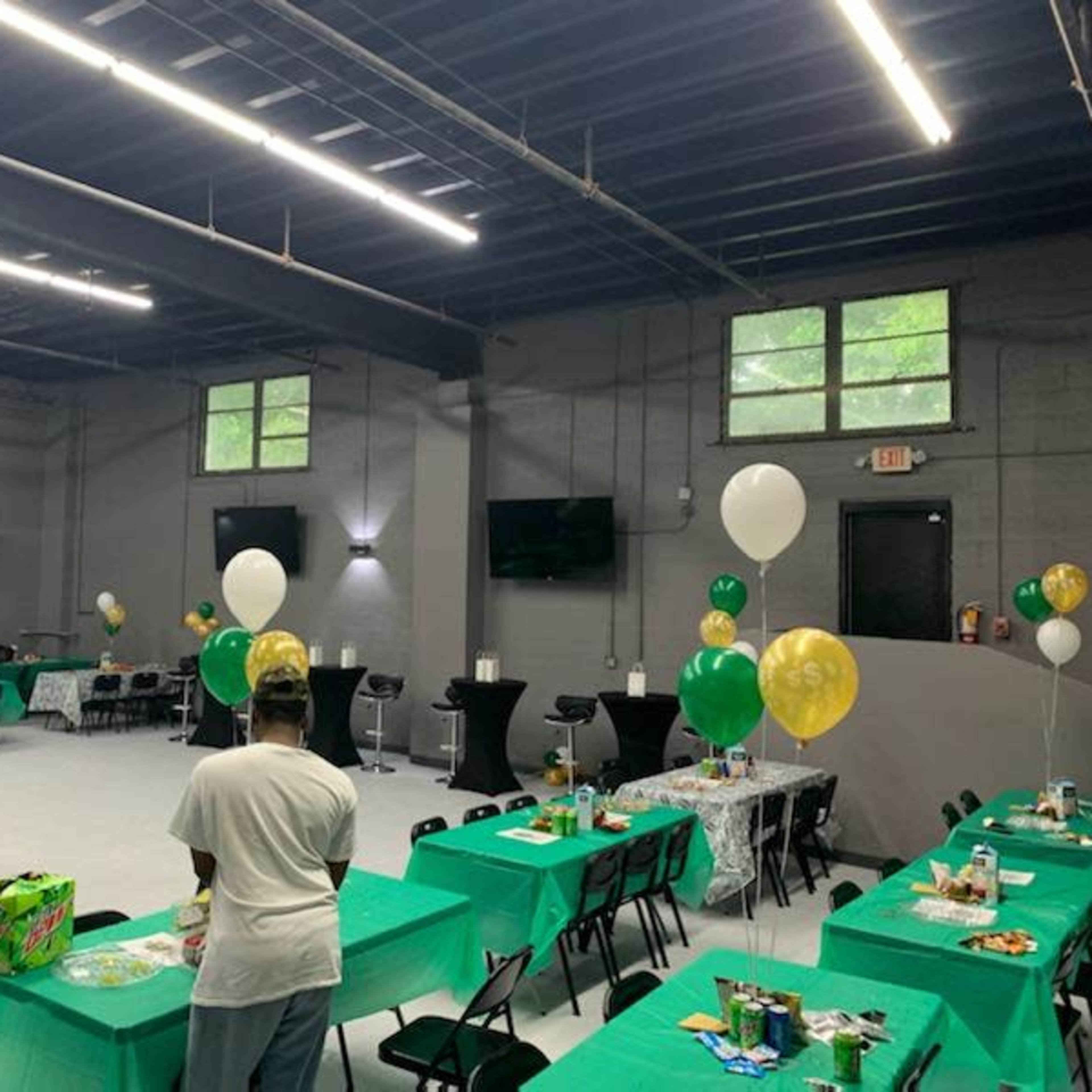 A spacious event room is set up with green and gold tablecloths, balloons, and various food items on tables for a gathering.