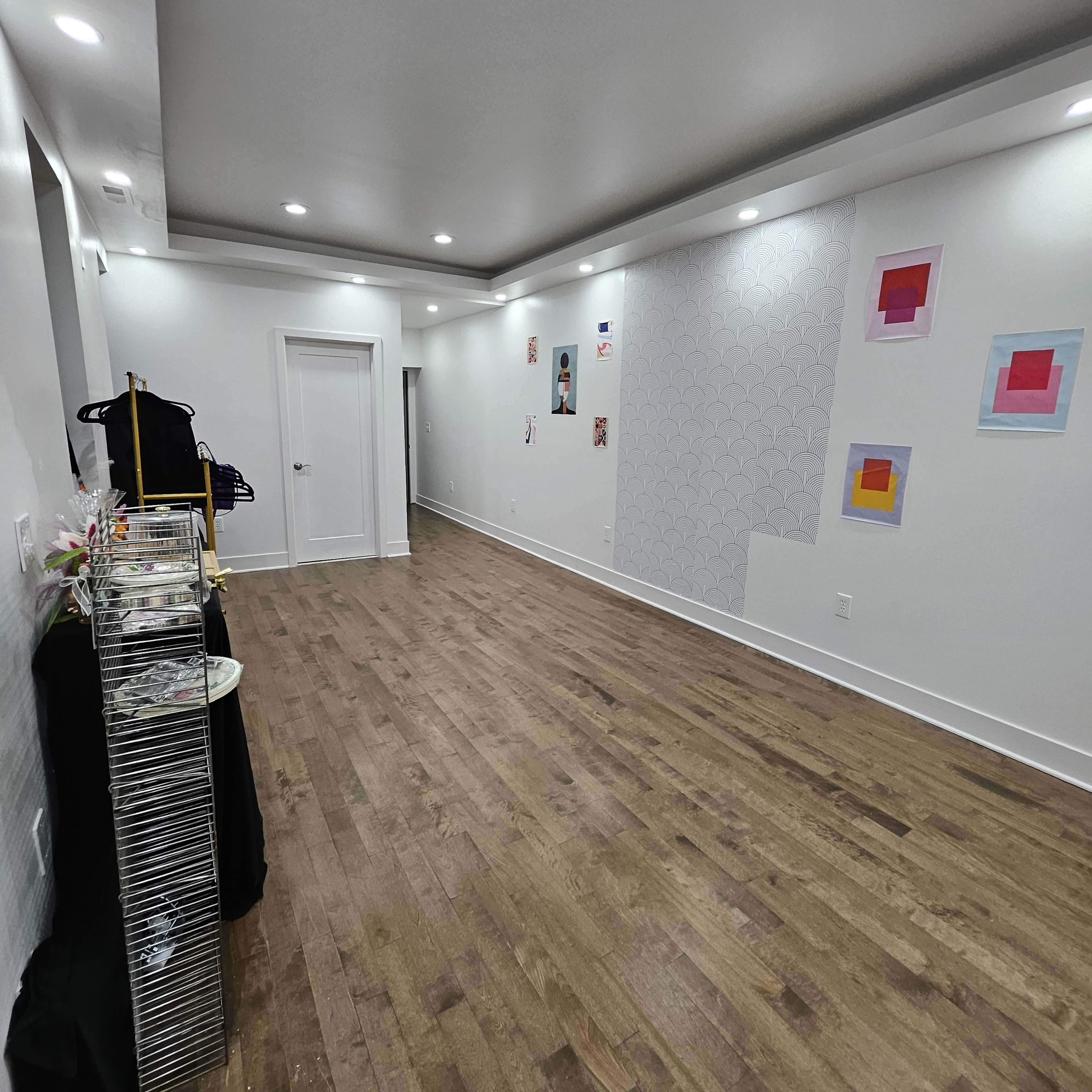 A spacious room with wooden flooring, white walls, and minimal decorations, featuring art pieces on the walls and a clothing rack on the left.