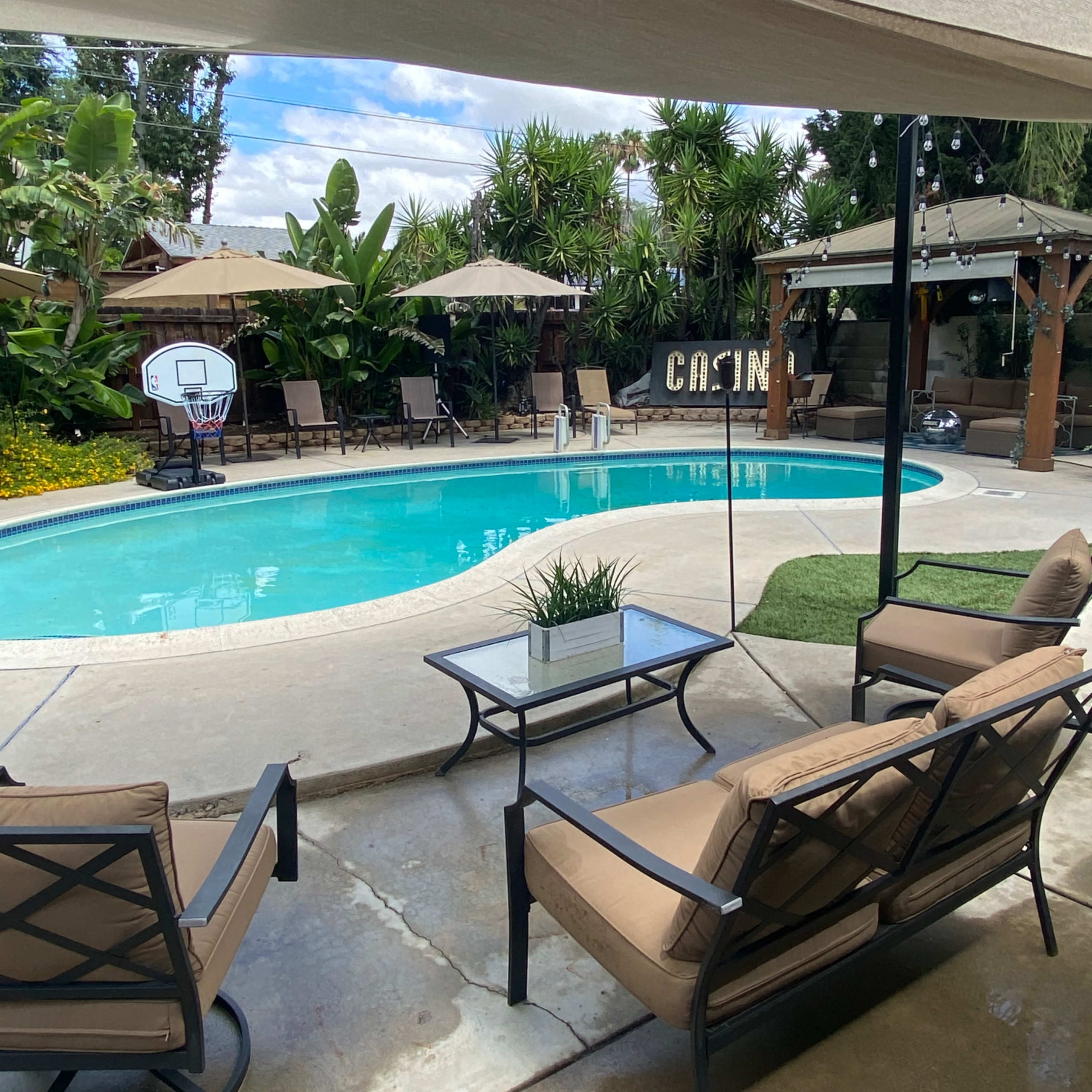 A seating area with two chairs and a glass table overlooks a swimming pool surrounded by palm trees and patio umbrellas.