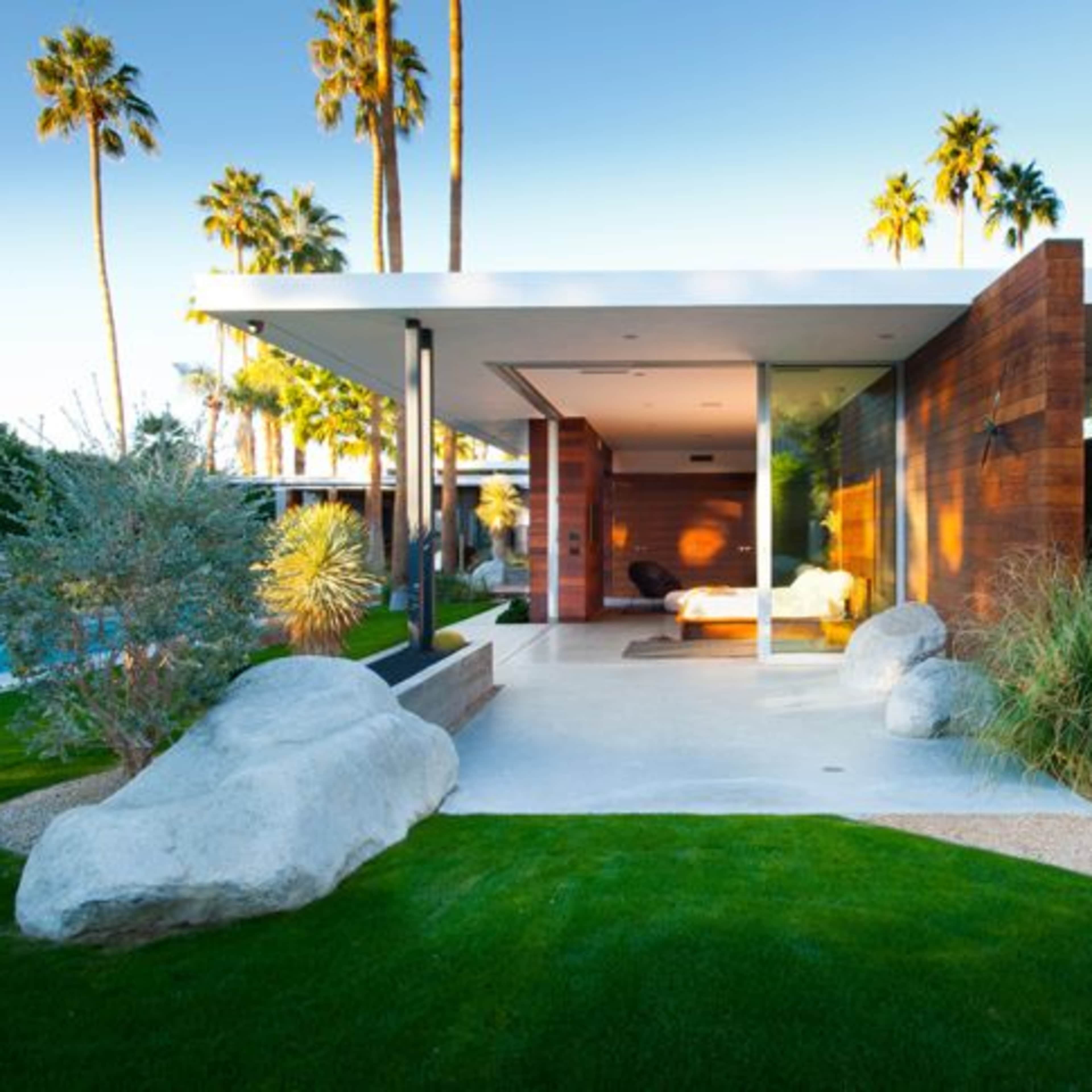 A modern single-story house with a flat roof is surrounded by lush greenery and large rocks, featuring a patio that opens to a swimming pool area.