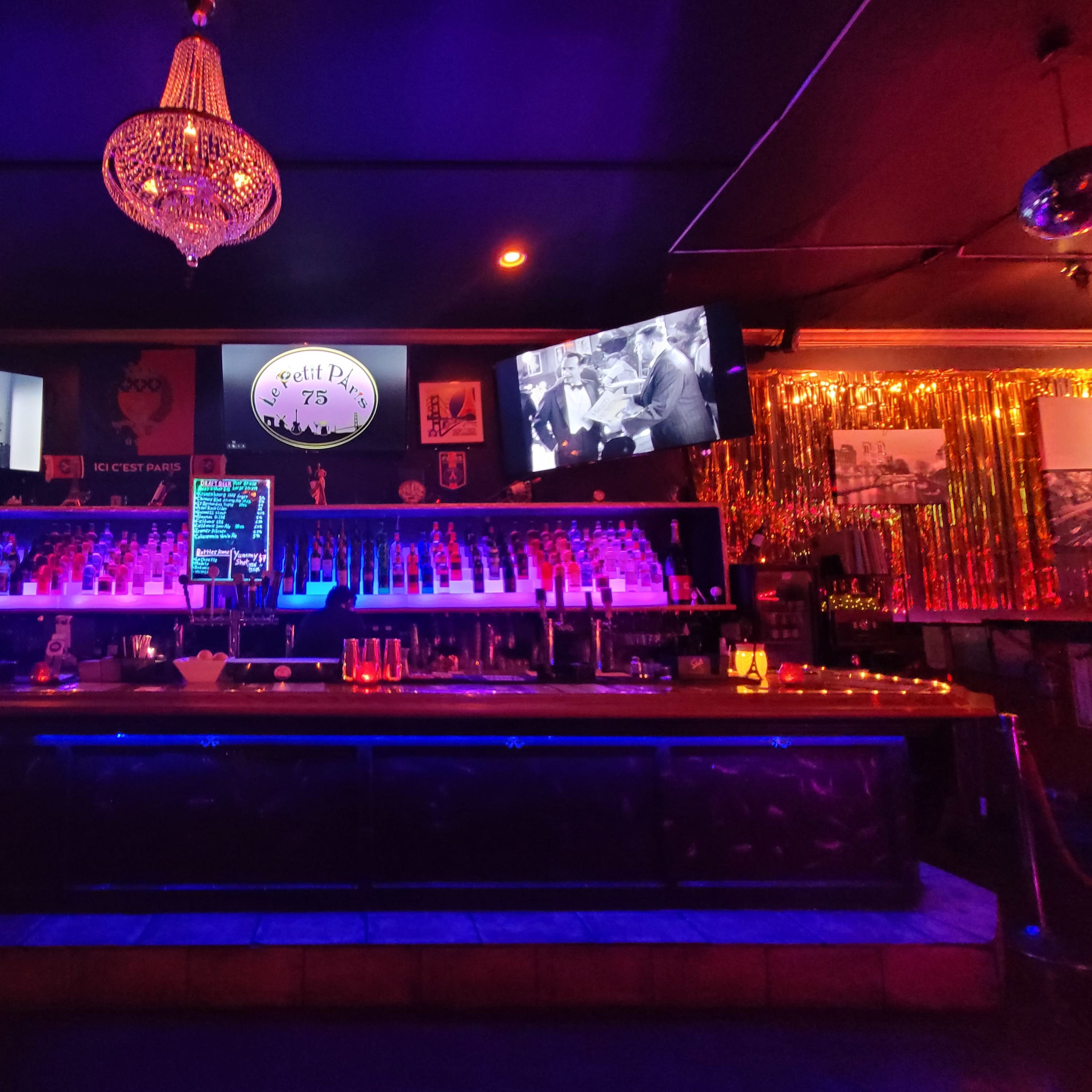 A dimly lit bar with two large screens displaying black-and-white footage, illuminated by colorful lights reflecting off a chandelier.