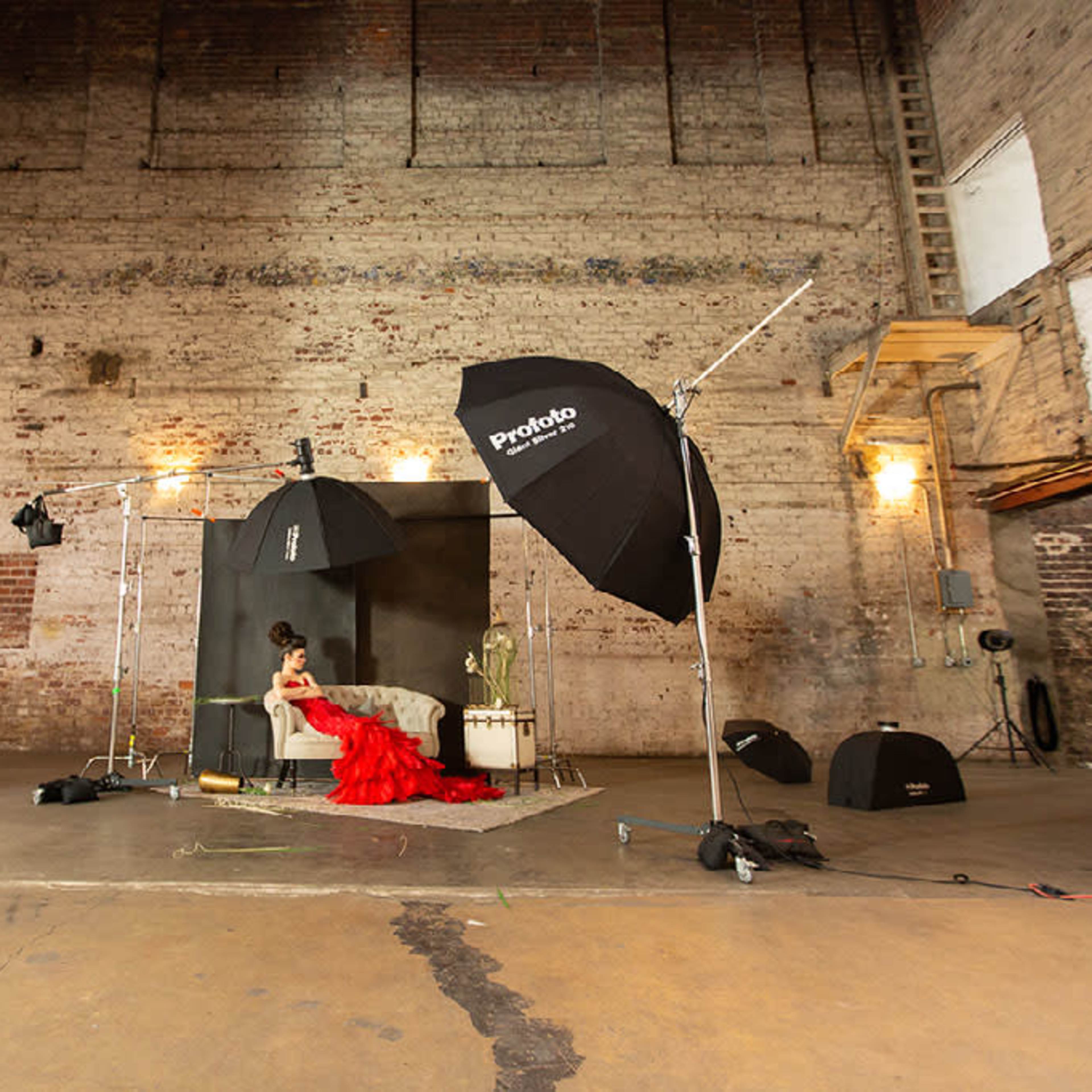 A woman in a red dress sits among photography equipment in an industrial studio with exposed brick walls.