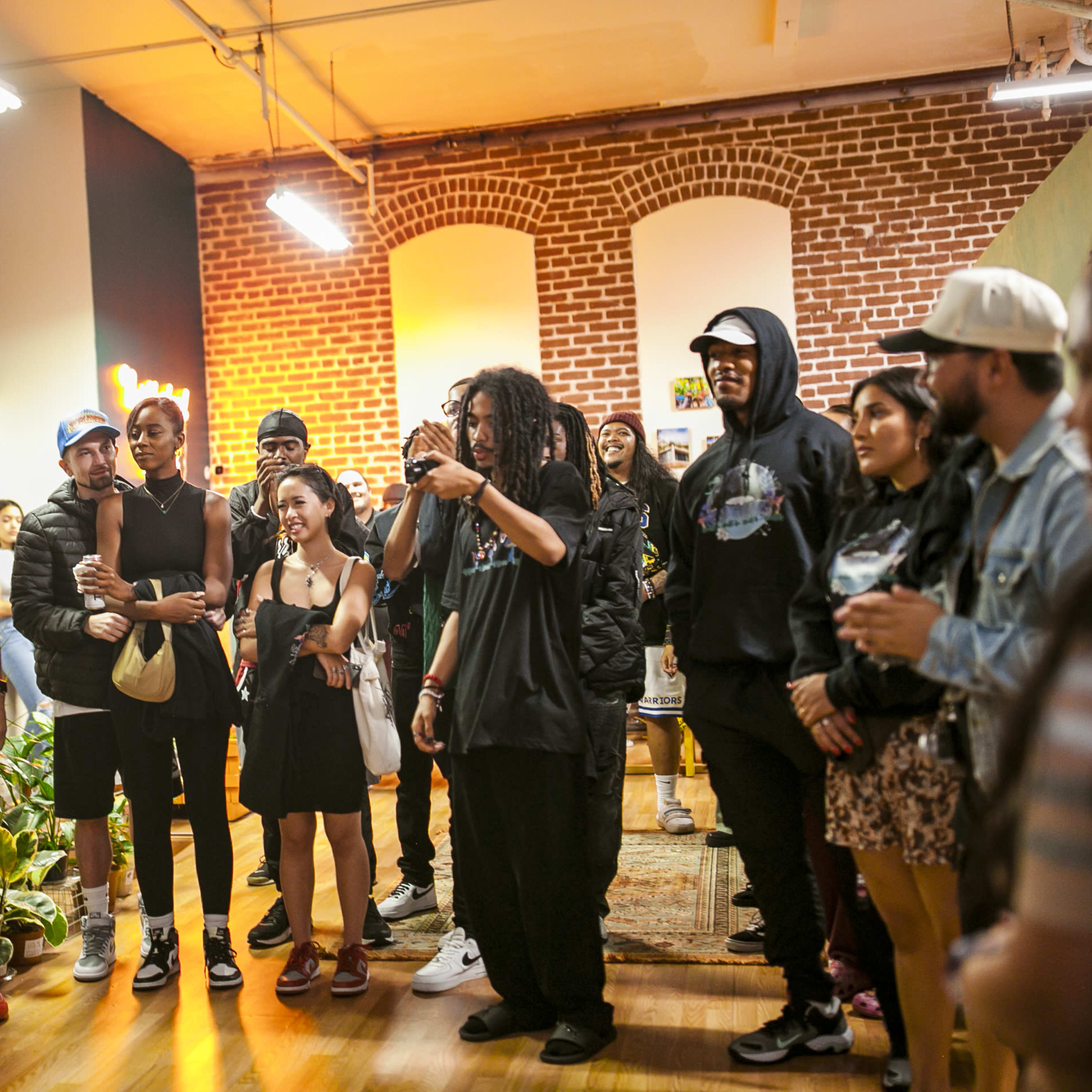A group of people gathers in a room with exposed brick walls, some holding phones and drinks while watching an event.