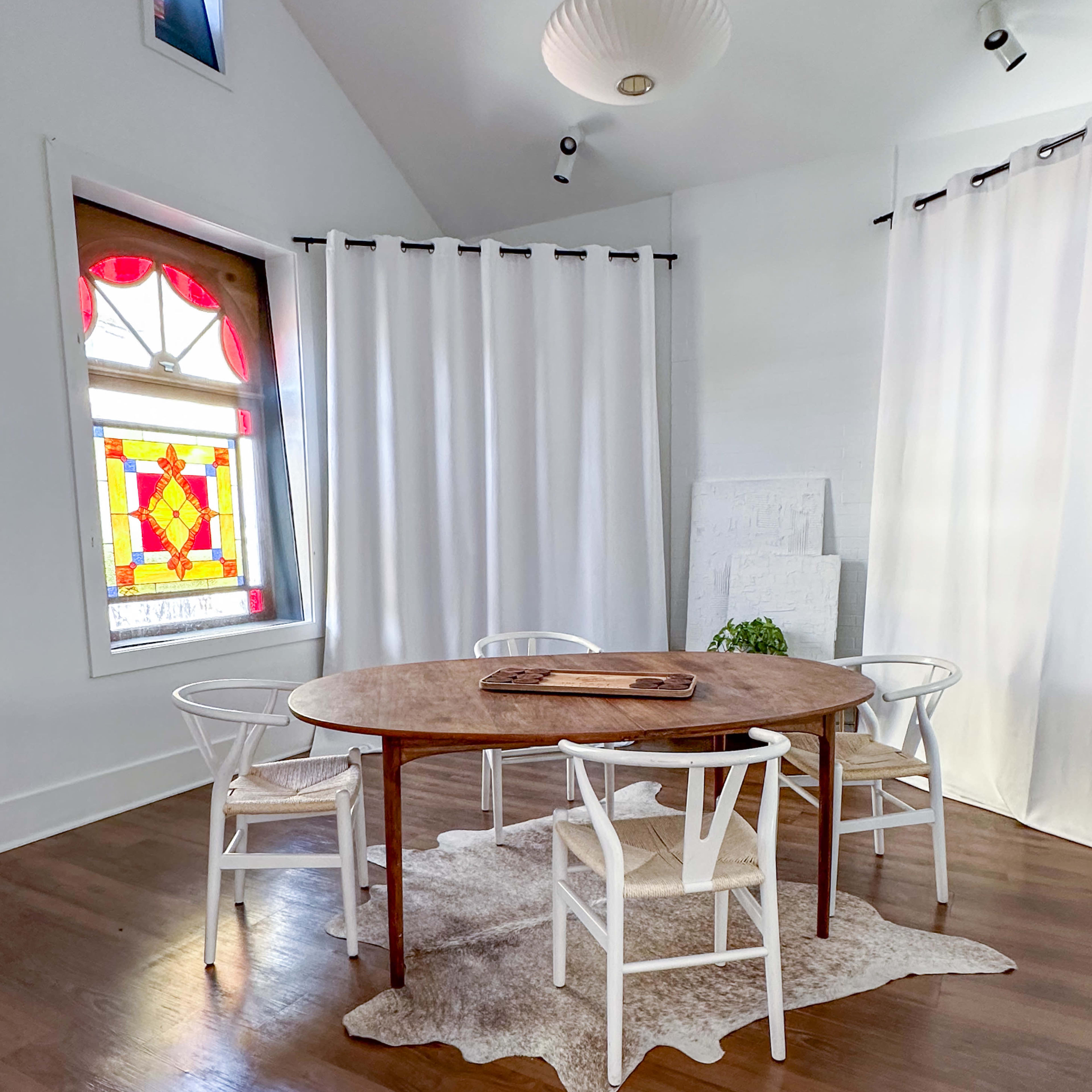 A round wooden table surrounded by white chairs sits on a light-colored rug in a bright room with stained glass windows and sheer curtains.