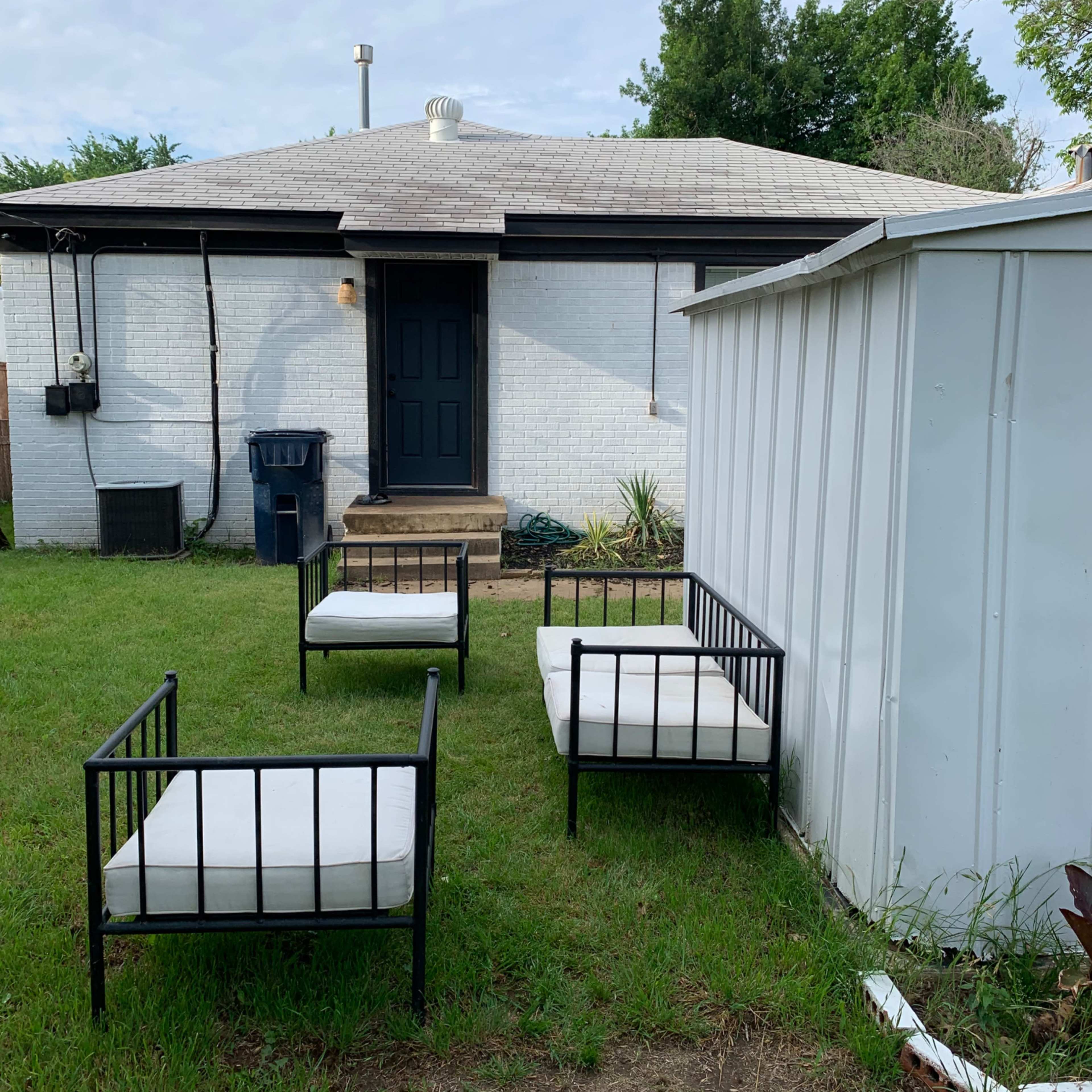 Three outdoor chairs are arranged on the grass in front of a white house with a dark door and a nearby storage shed.