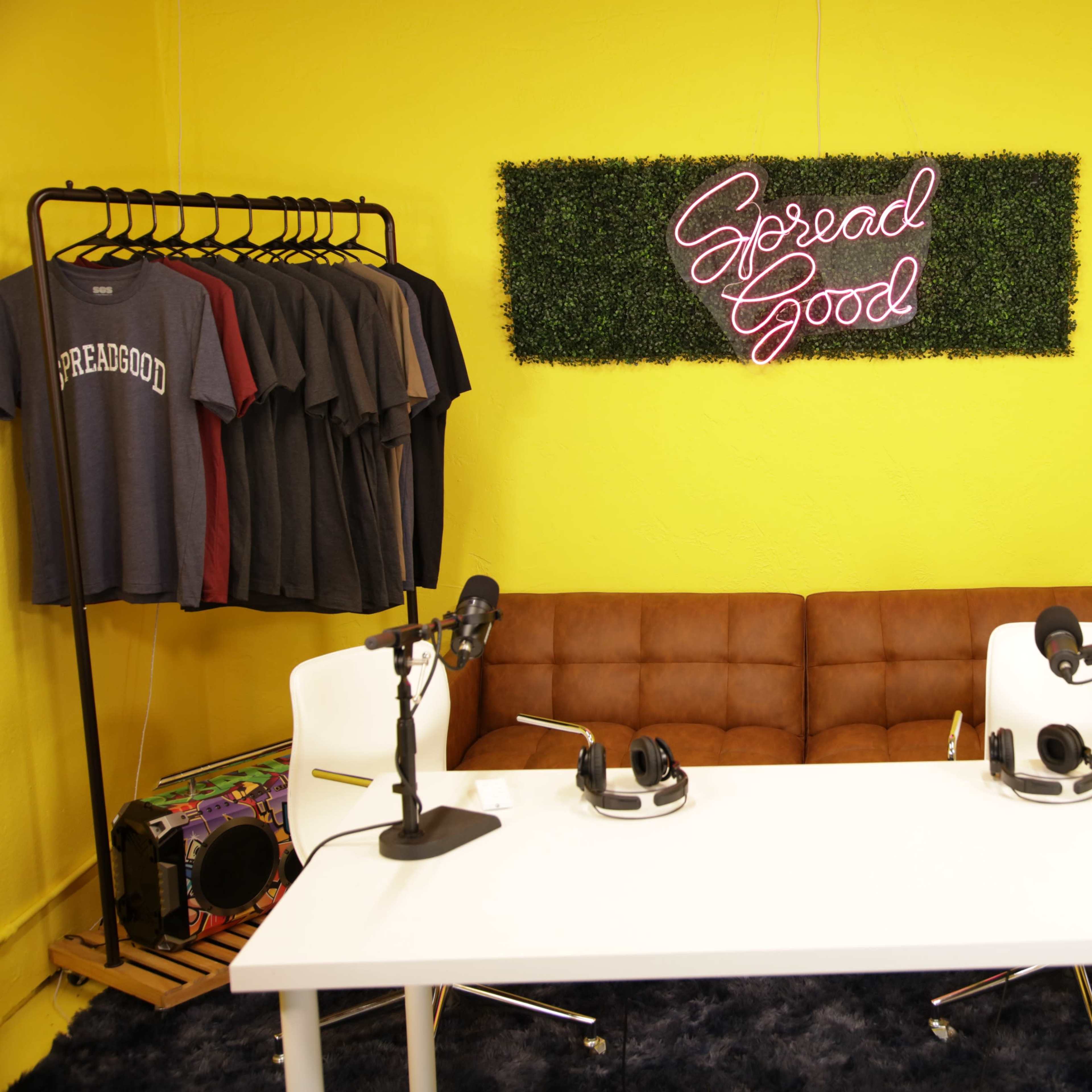 A recording studio setup features a white table with two microphones and headphones, a brown couch, a clothing rack with t-shirts, and a neon sign on a yellow wall that reads "Spread Good."