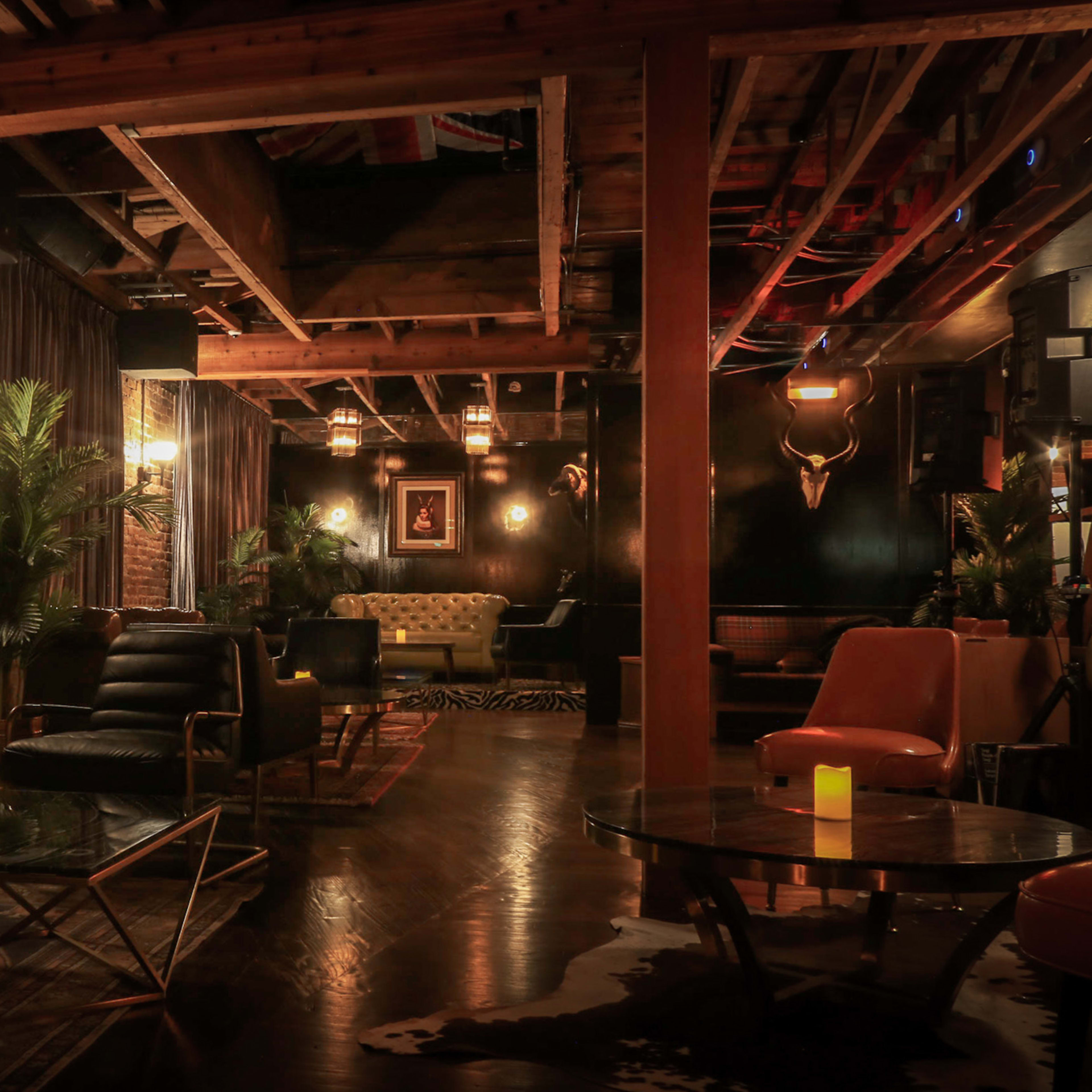 A dimly lit lounge featuring leather seating, wooden beams, brick walls, and potted plants, with candles on low tables.