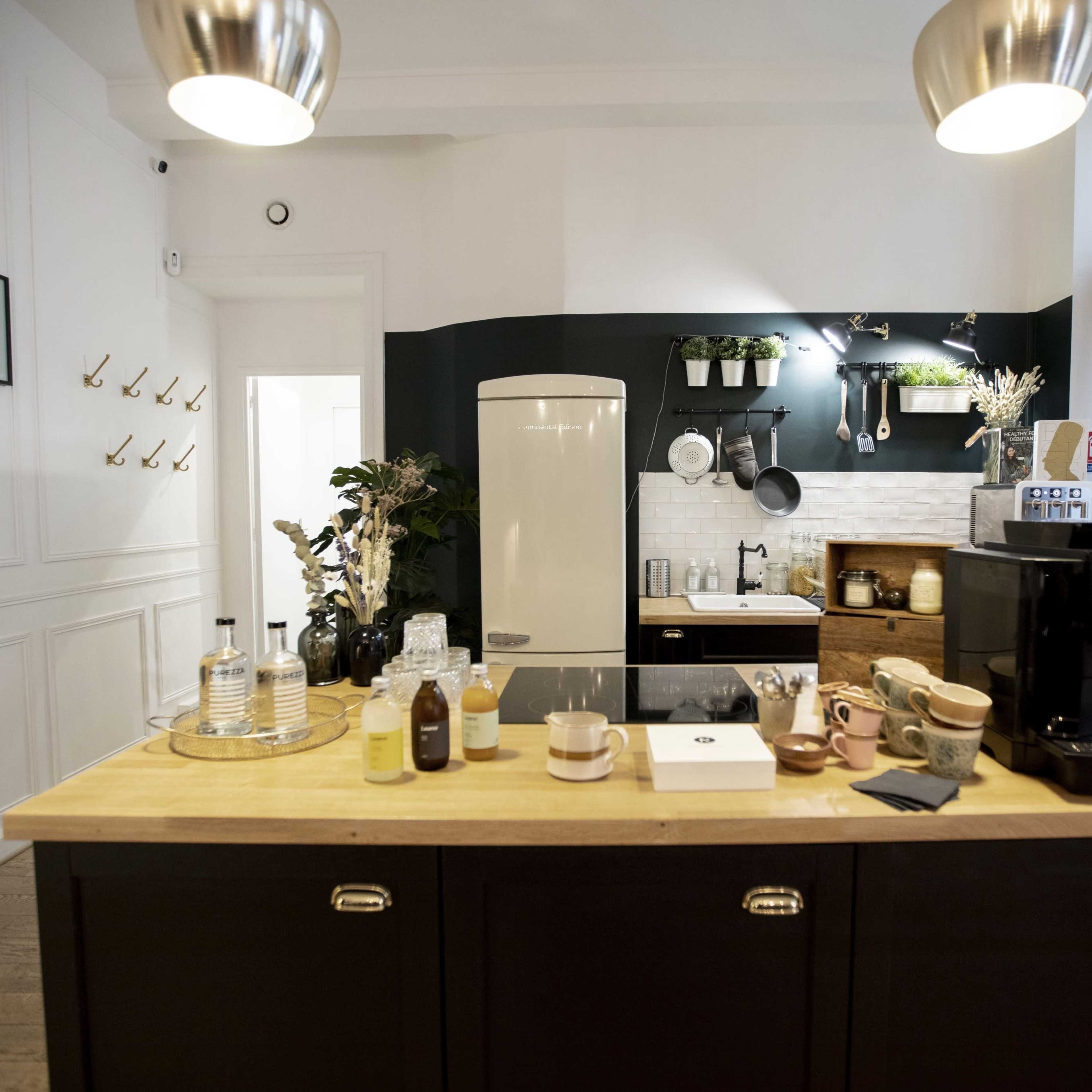 A modern kitchen with a central wooden island, featuring various kitchen appliances, utensils, and decorative plants, set against a backdrop of white walls and dark green accents.