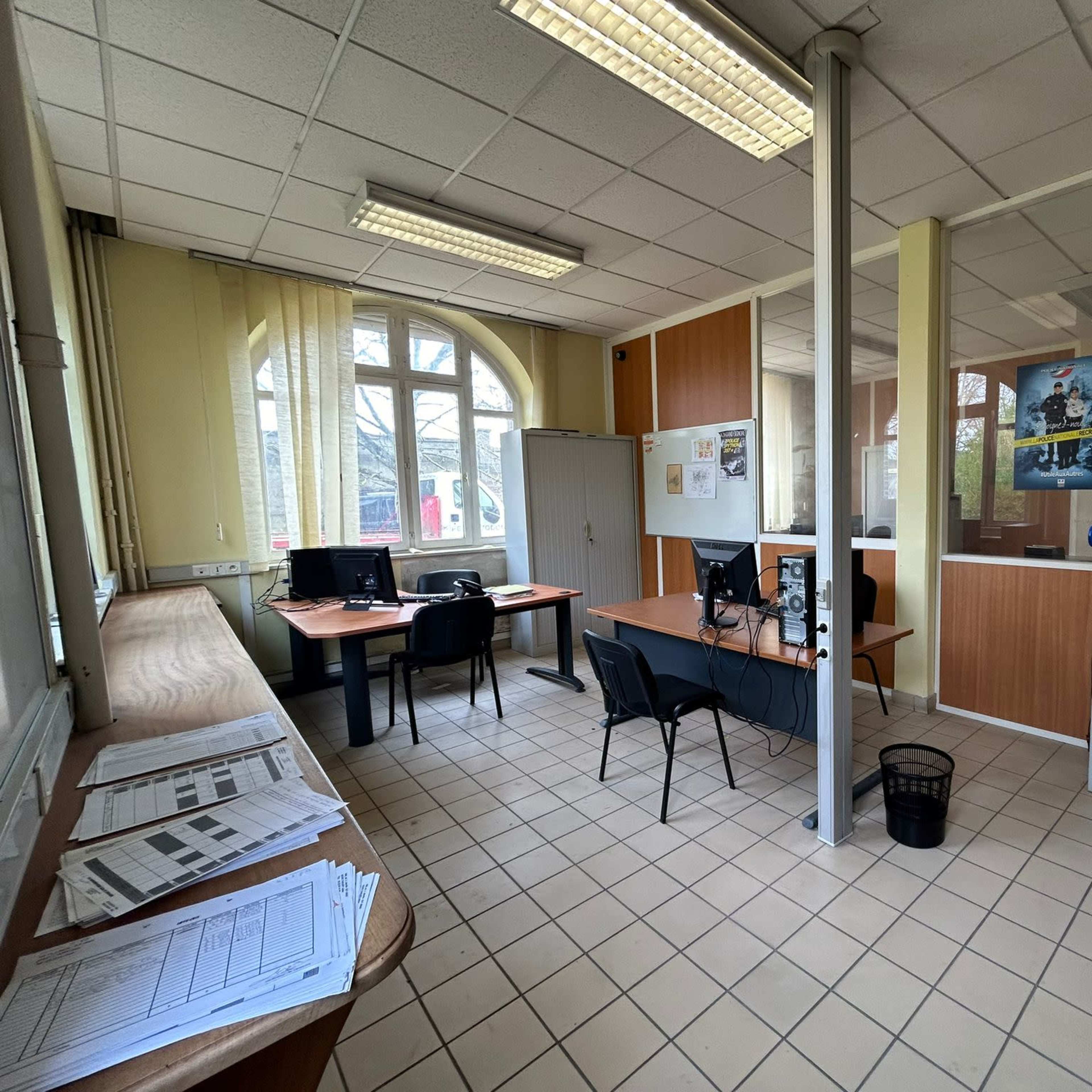 A modest office space with several desks, computers, filing cabinets, and large windows allowing natural light.