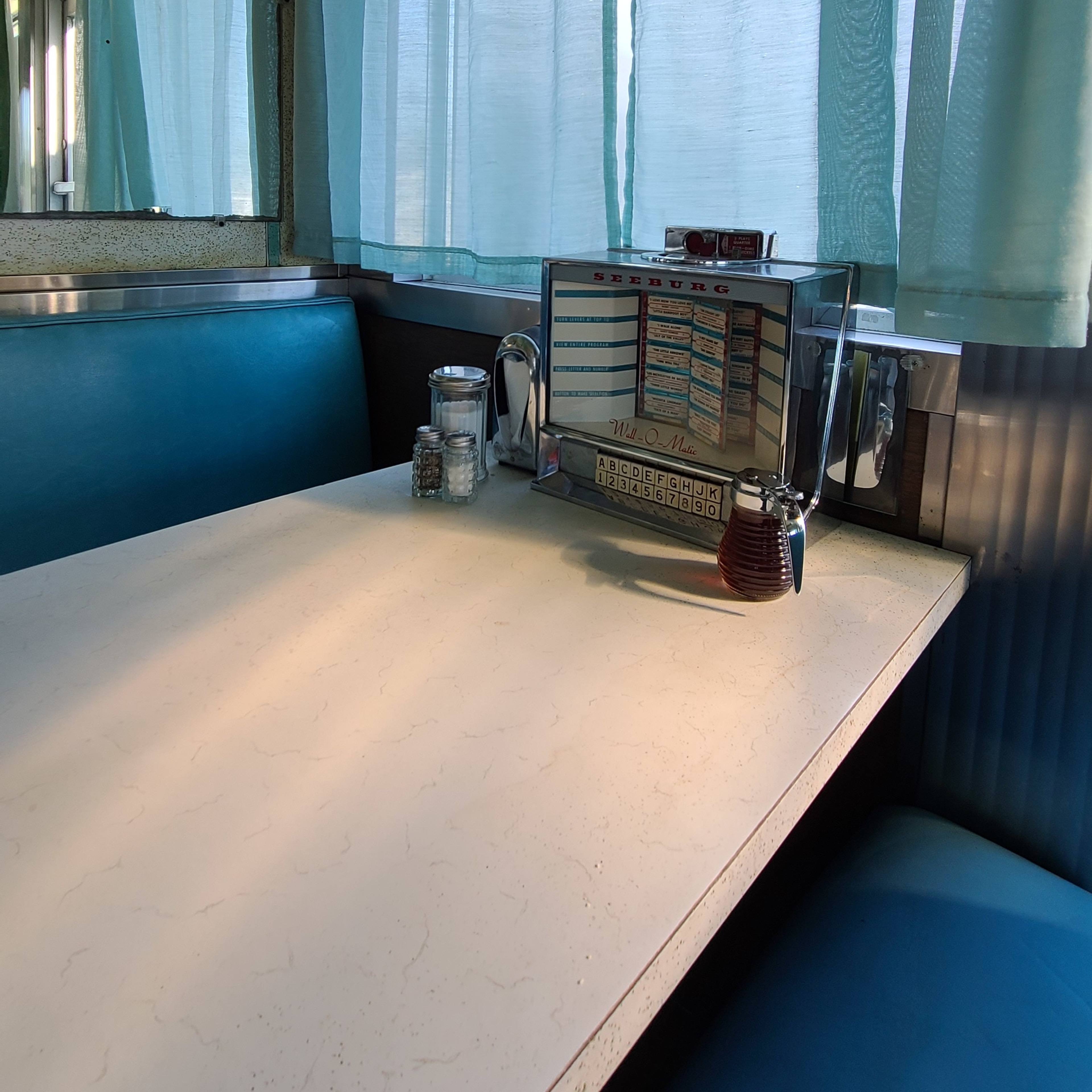 A diner booth with a jukebox and condiments on a table, surrounded by light teal curtains.