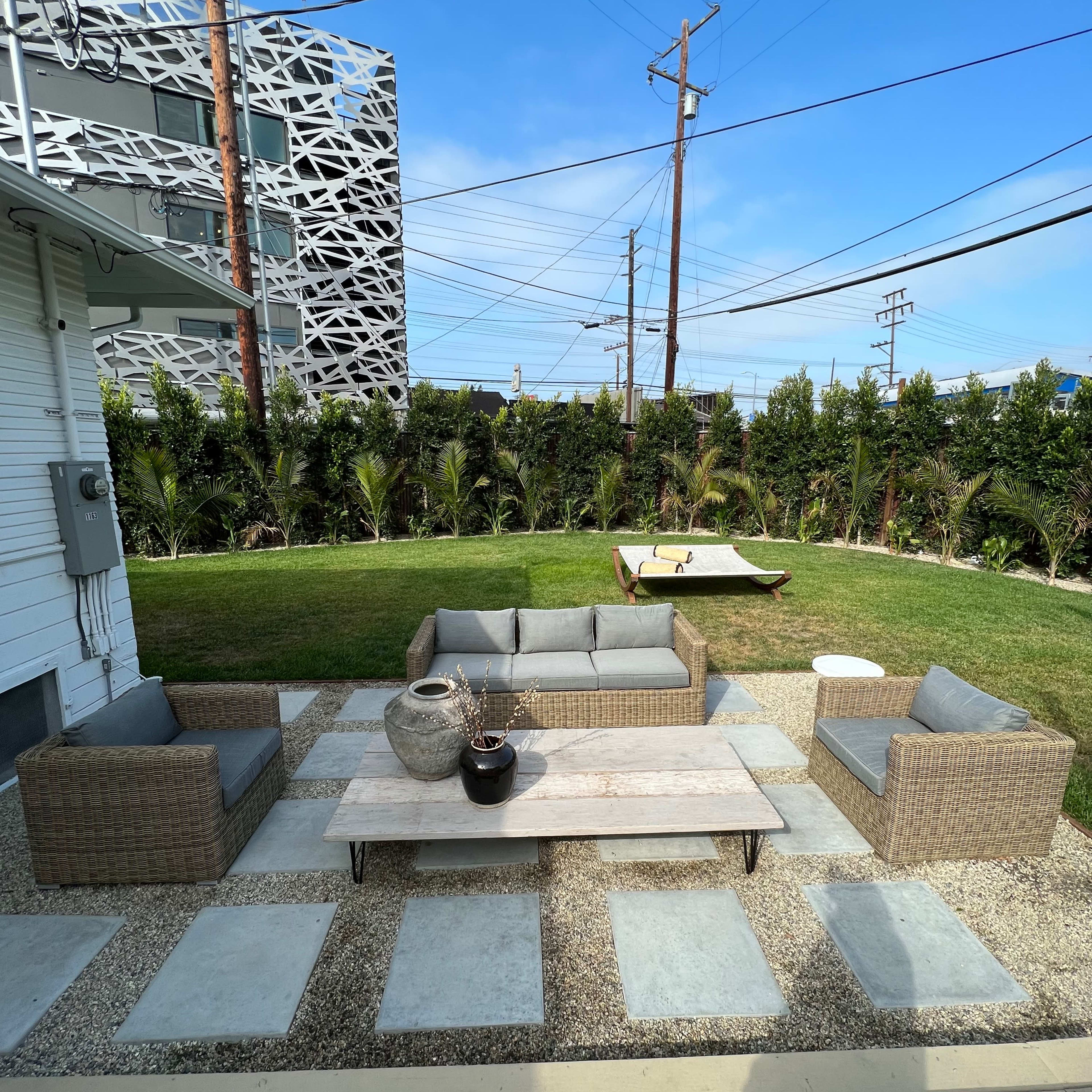 A patio area features a gray sectional sofa set and a wooden coffee table, surrounded by stone pavers and a green lawn with palm trees in the background.