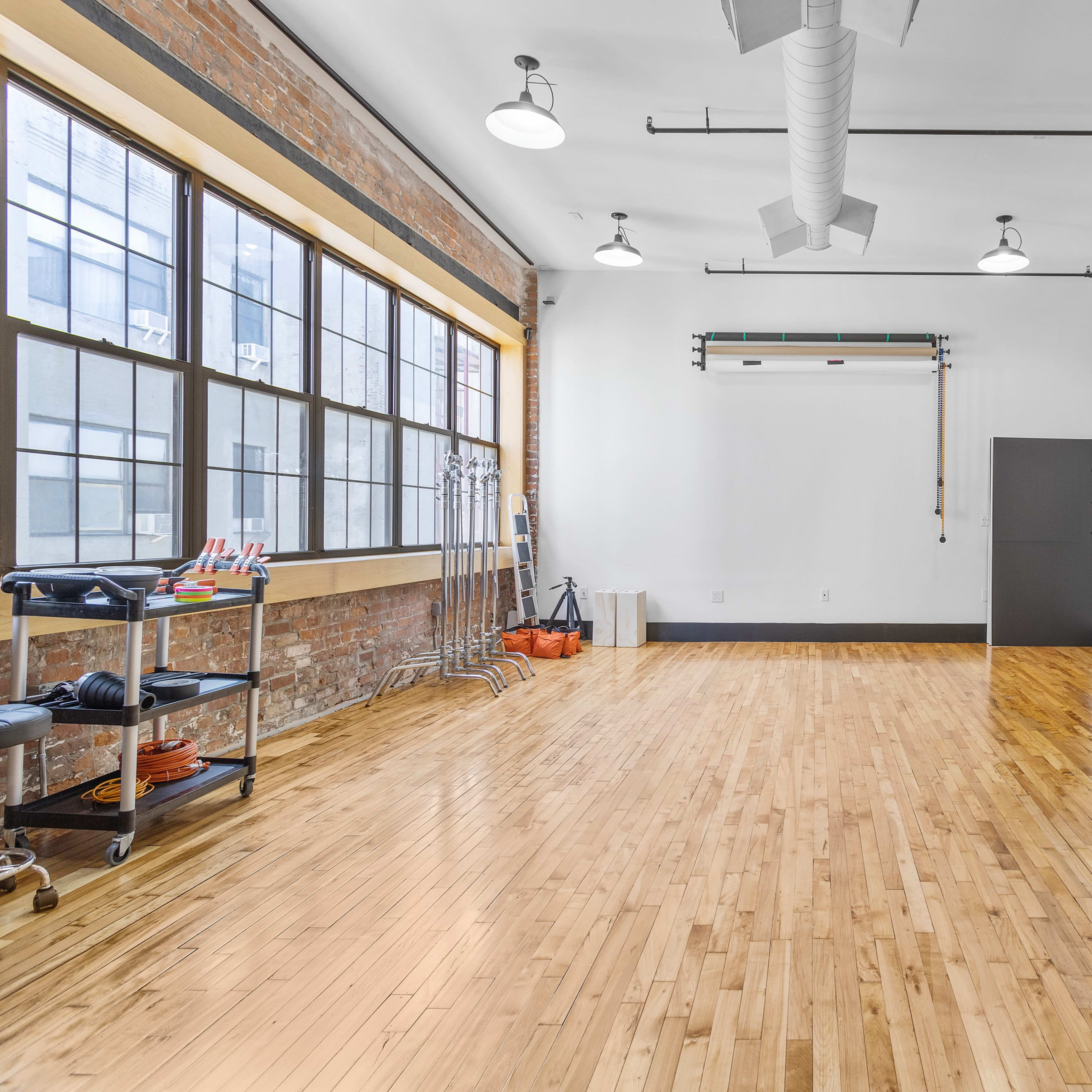 A spacious, minimally decorated studio with large windows, wooden flooring, and a vanity mirror on a table against a white wall.