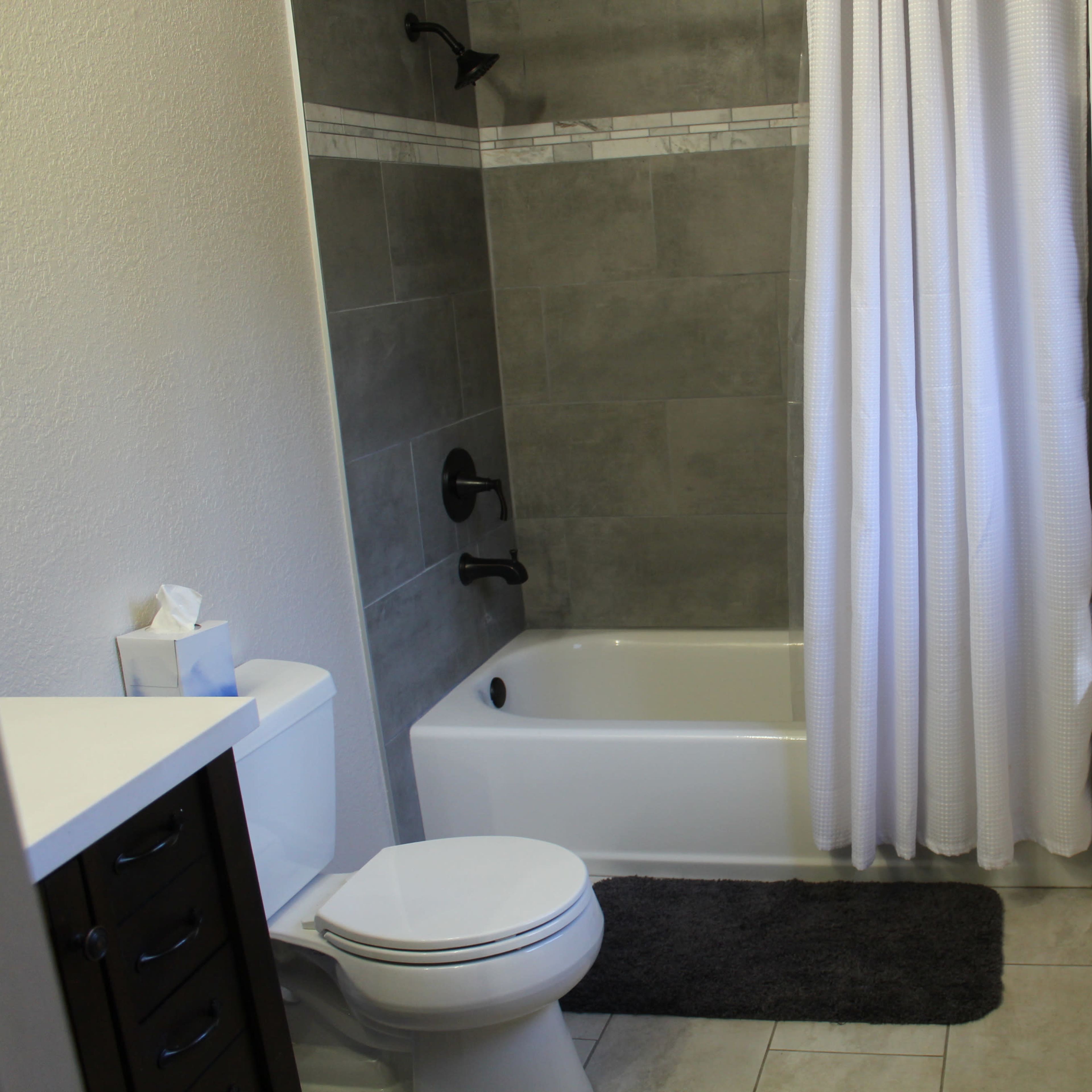 A bathroom with a shower-tub combination, a toilet, a sink with a dark vanity, and white curtains beside a window.