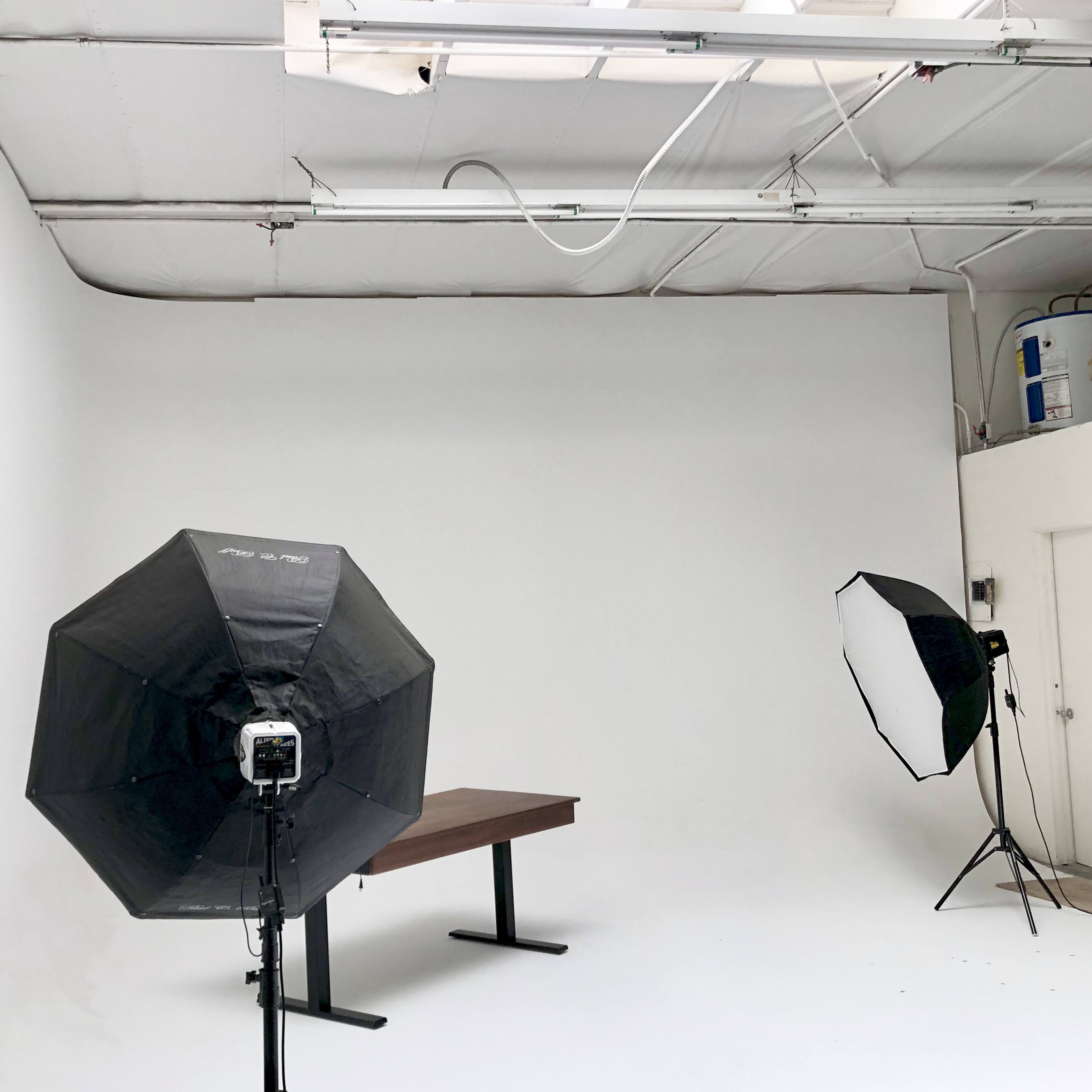 A photography studio with a plain white backdrop, a wooden table in the center, and two lighting setups positioned on either side.