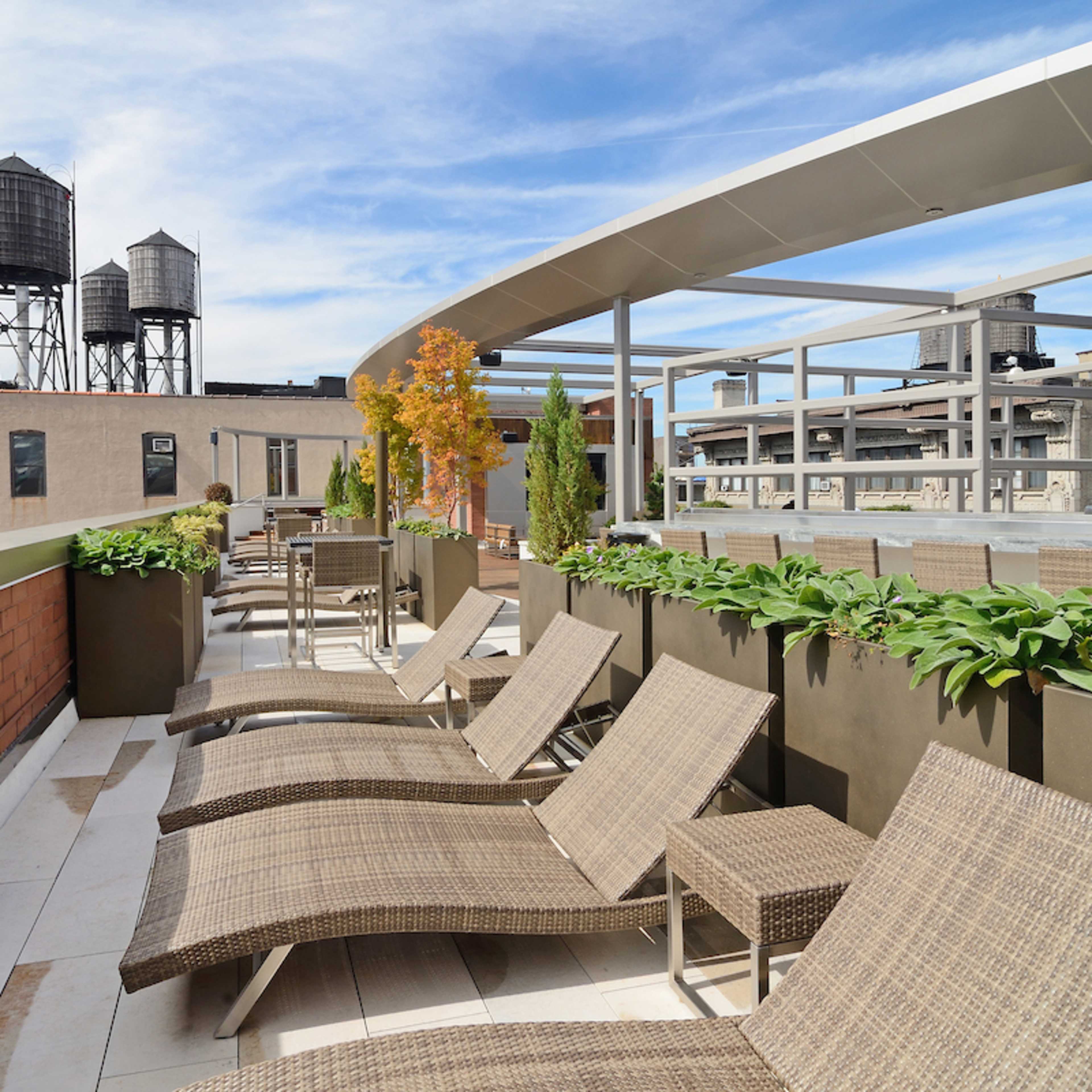 A rooftop terrace with lounge chairs, potted plants, and water towers in the background under a blue sky.