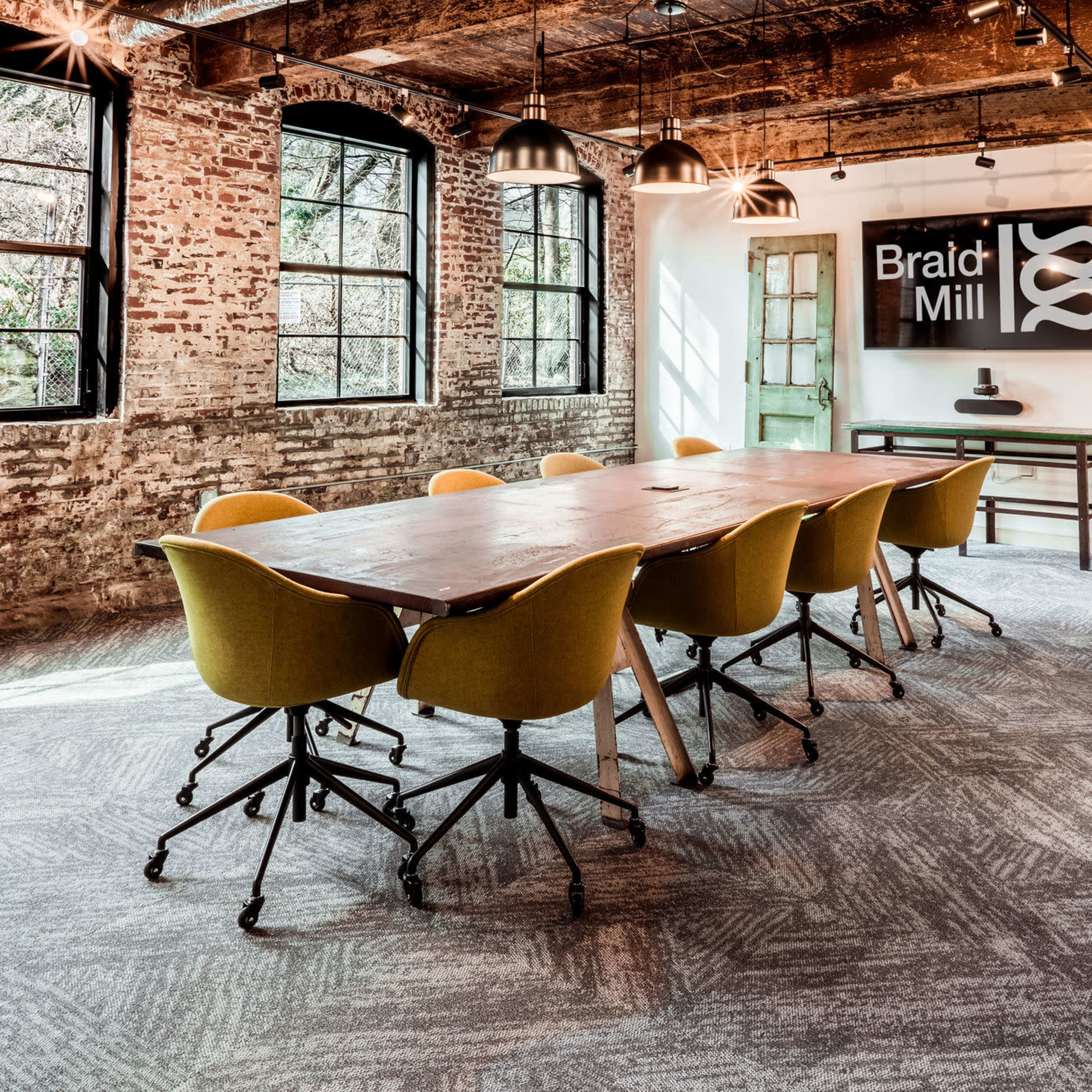 A modern meeting room featuring a large wooden table surrounded by yellow chairs, with exposed brick walls and large windows allowing natural light.