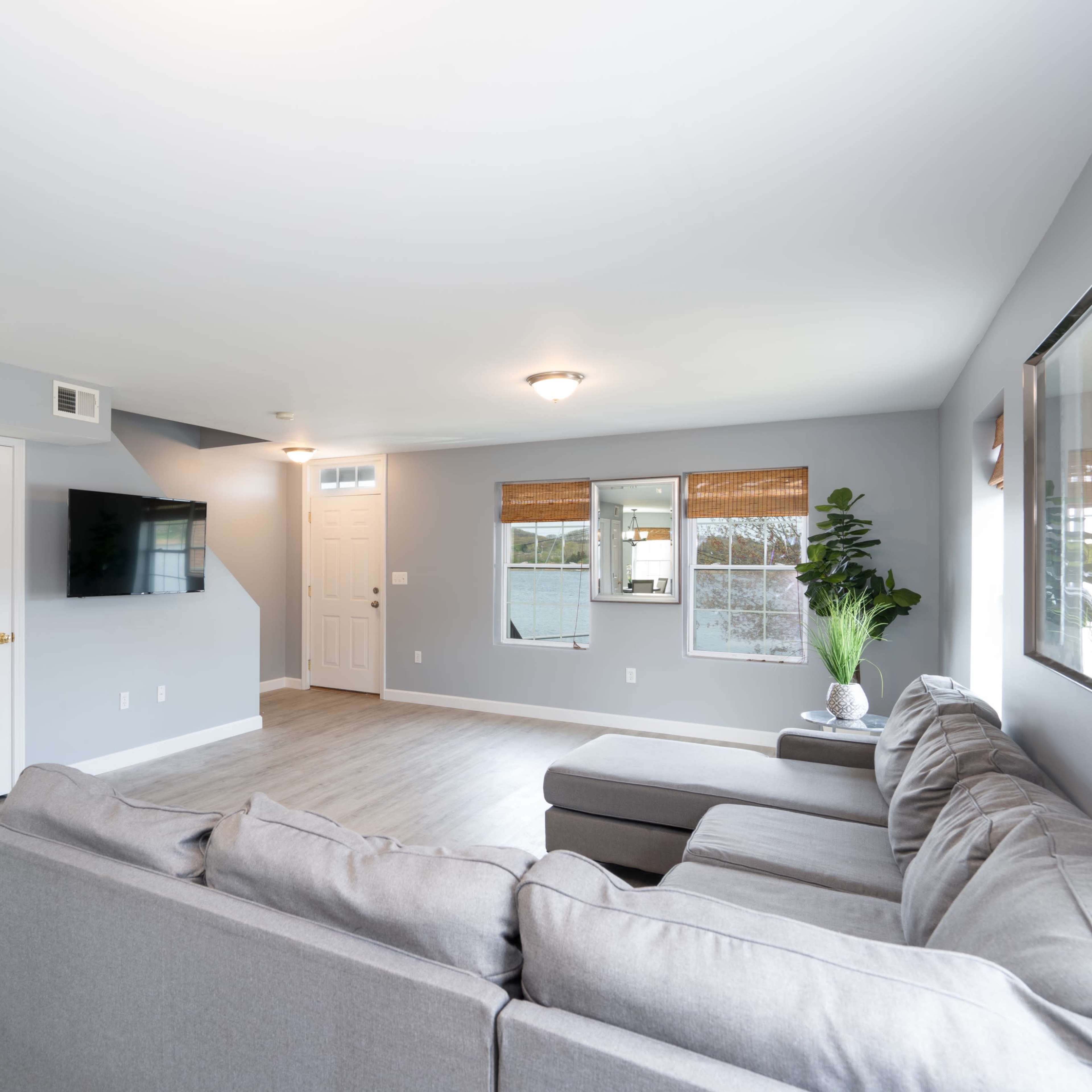 A modern living room with gray walls, a sectional sofa, a mounted television, and two windows with woven shades.