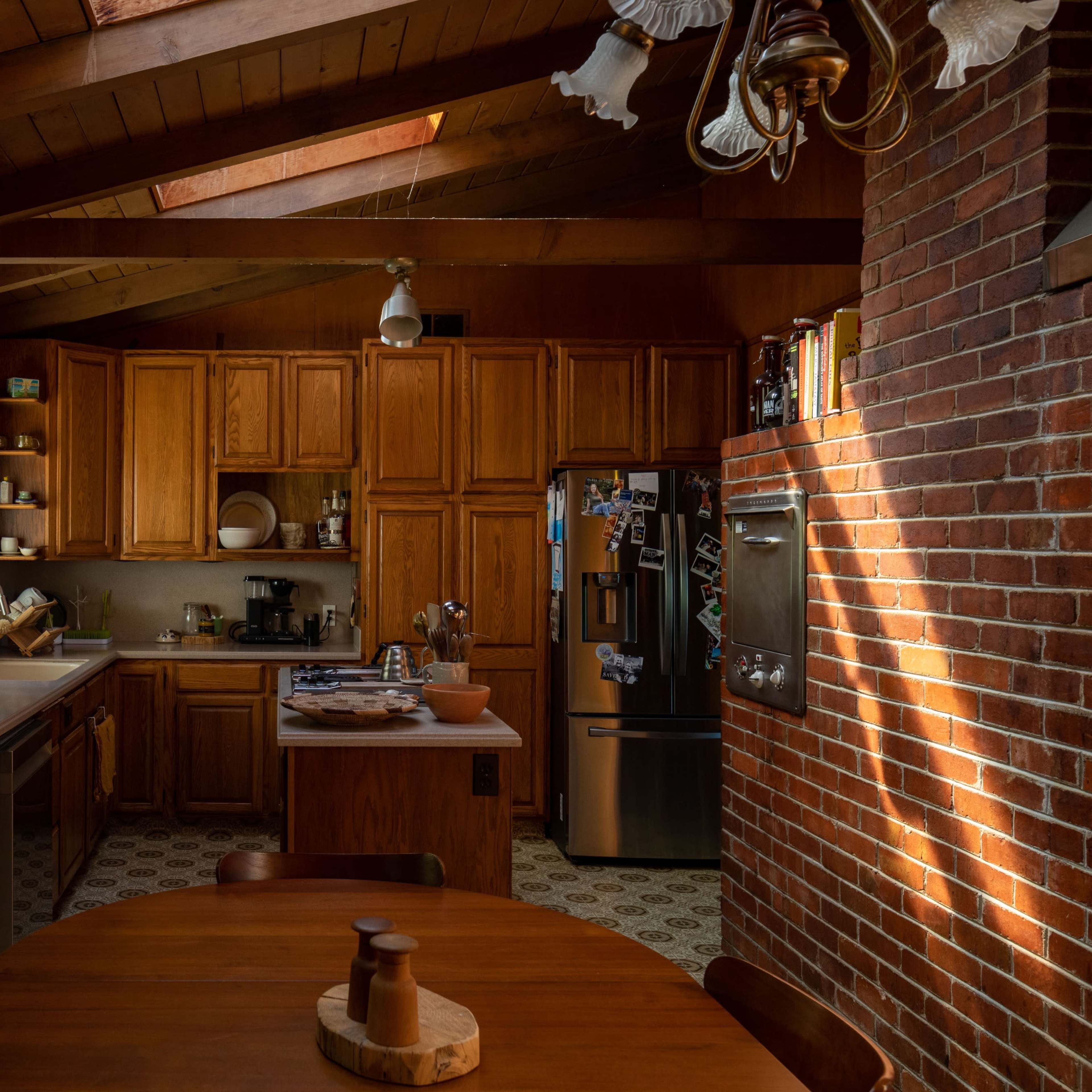 A wooden kitchen with a brick wall, featuring cabinets, a refrigerator, and a dining table in the foreground.