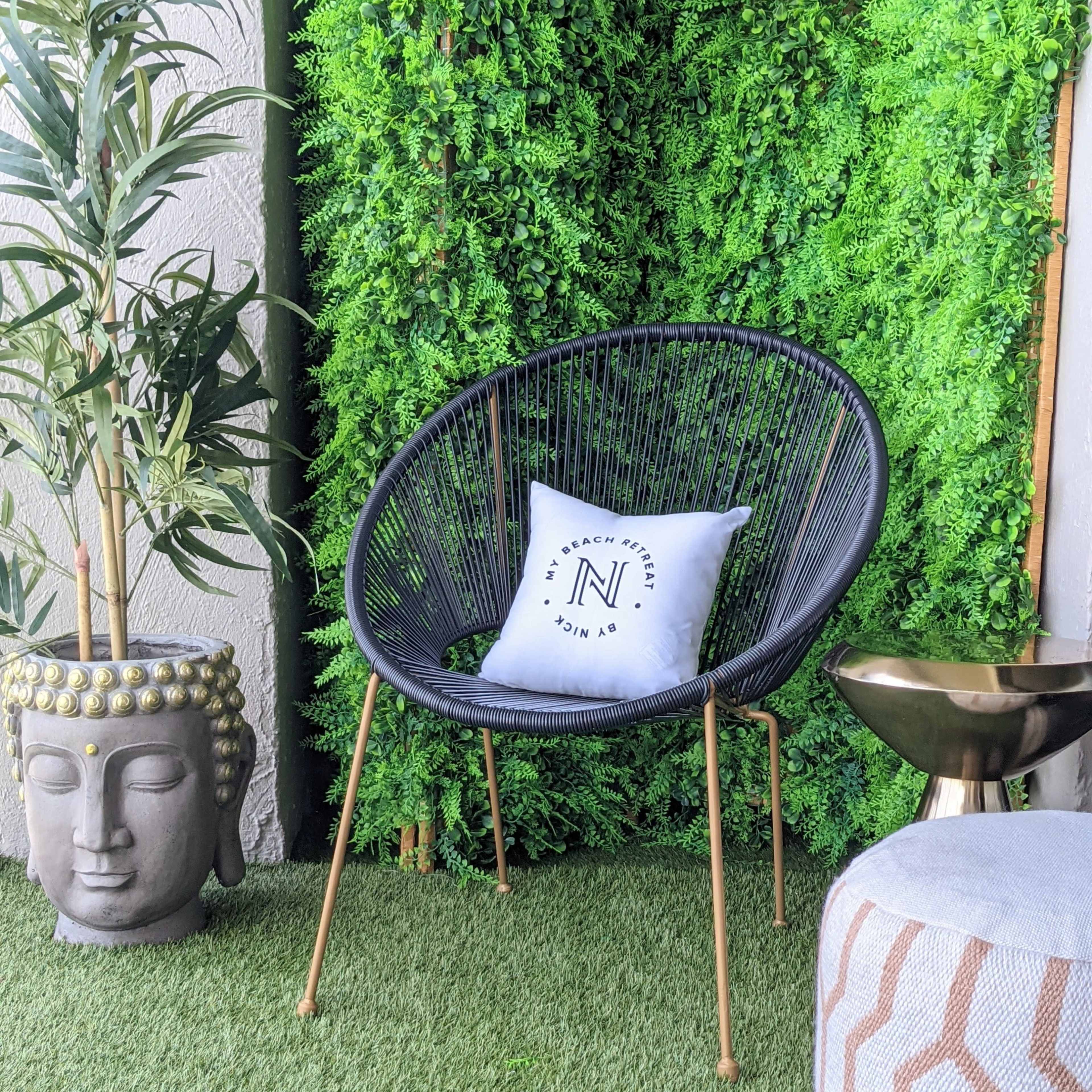 A black round chair with a decorative pillow, positioned next to a stone Buddha statue and a metallic side table, all against a backdrop of lush greenery.