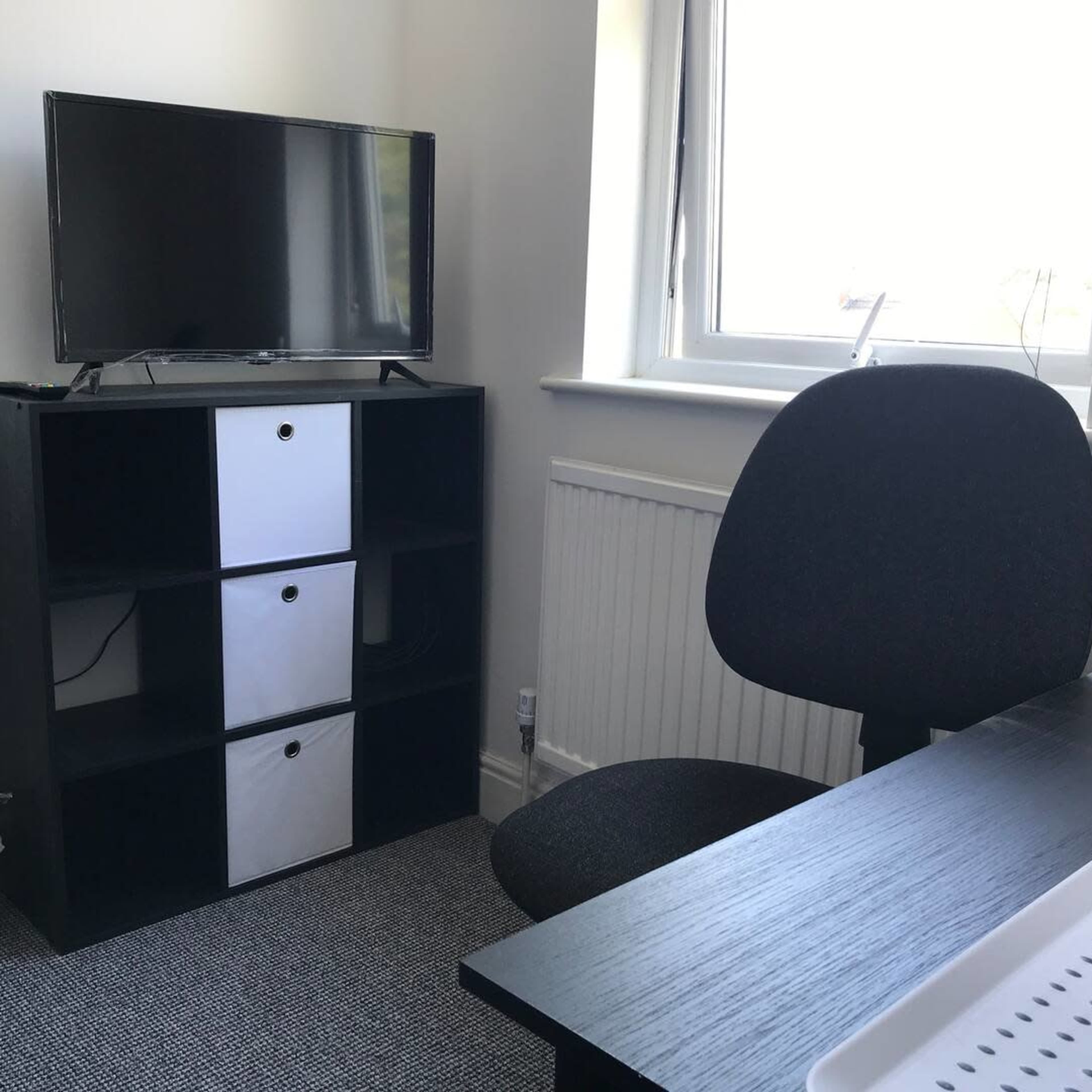 A room with a television on a black shelf, a gray chair, and a small white trash bin beside a radiator.