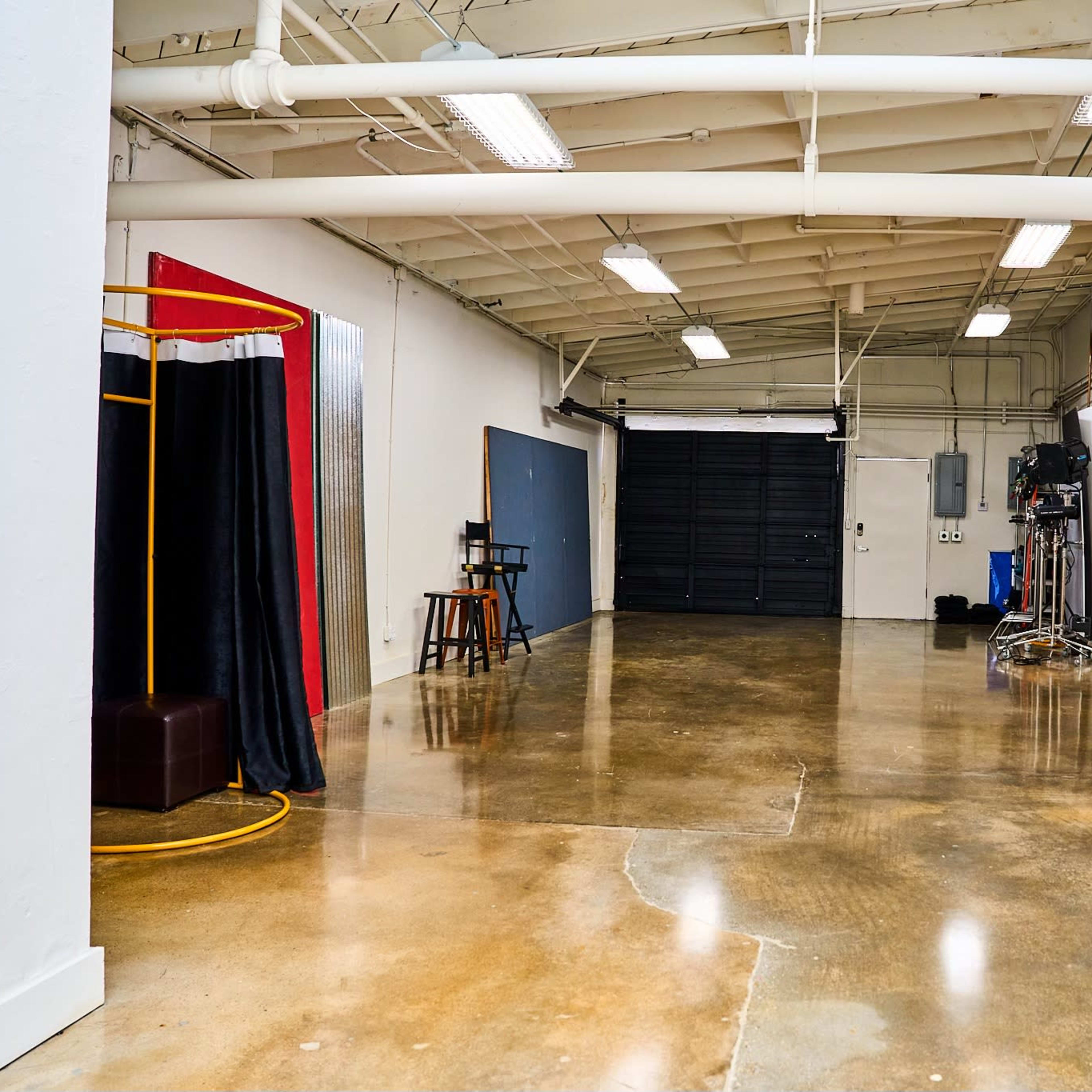 An empty interior space with polished concrete floors, featuring a black roll-up garage door, a mirrored table with chairs, and a photography backdrop setup in one corner.