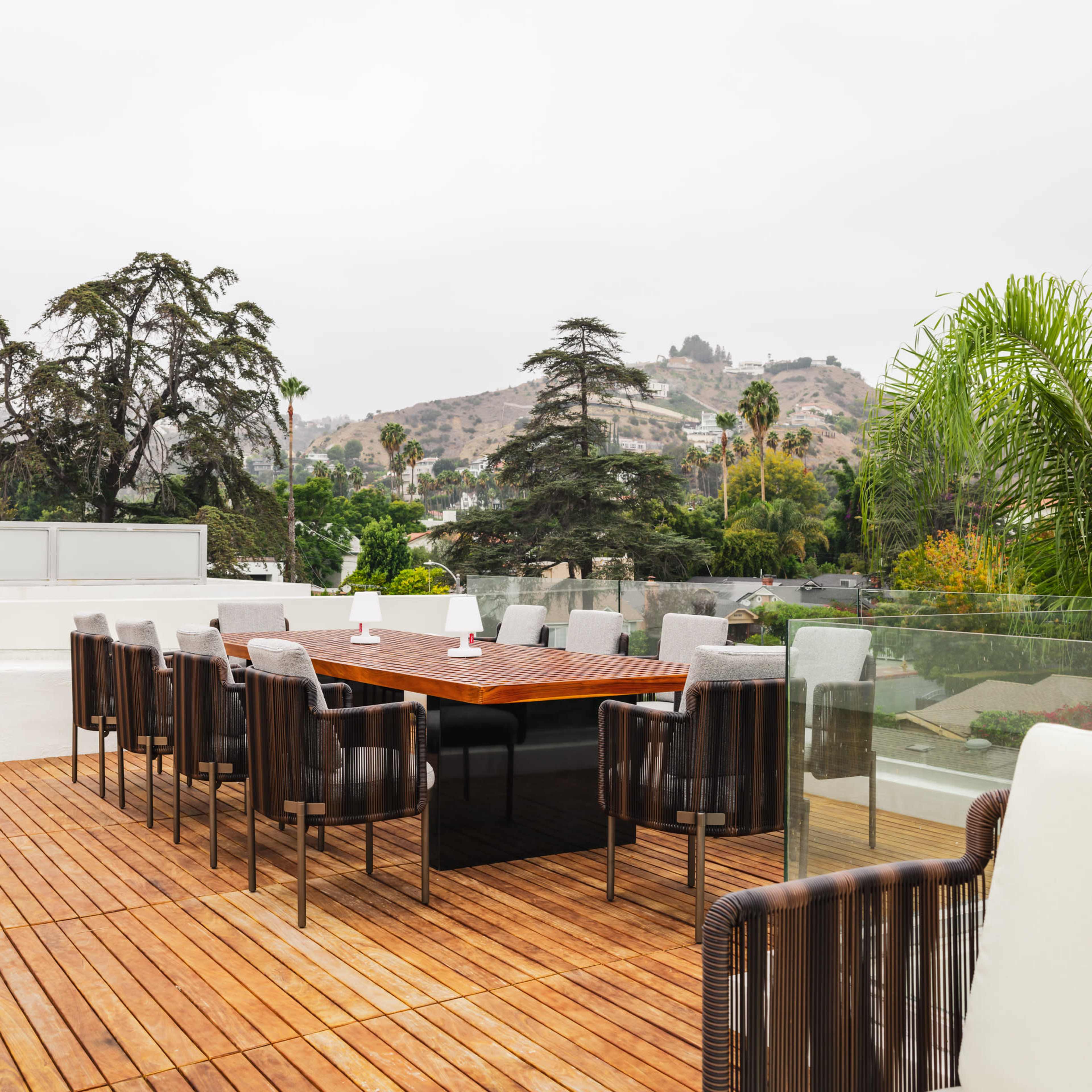 A spacious rooftop terrace features a large wooden dining table surrounded by chairs, set against a backdrop of hills and trees.