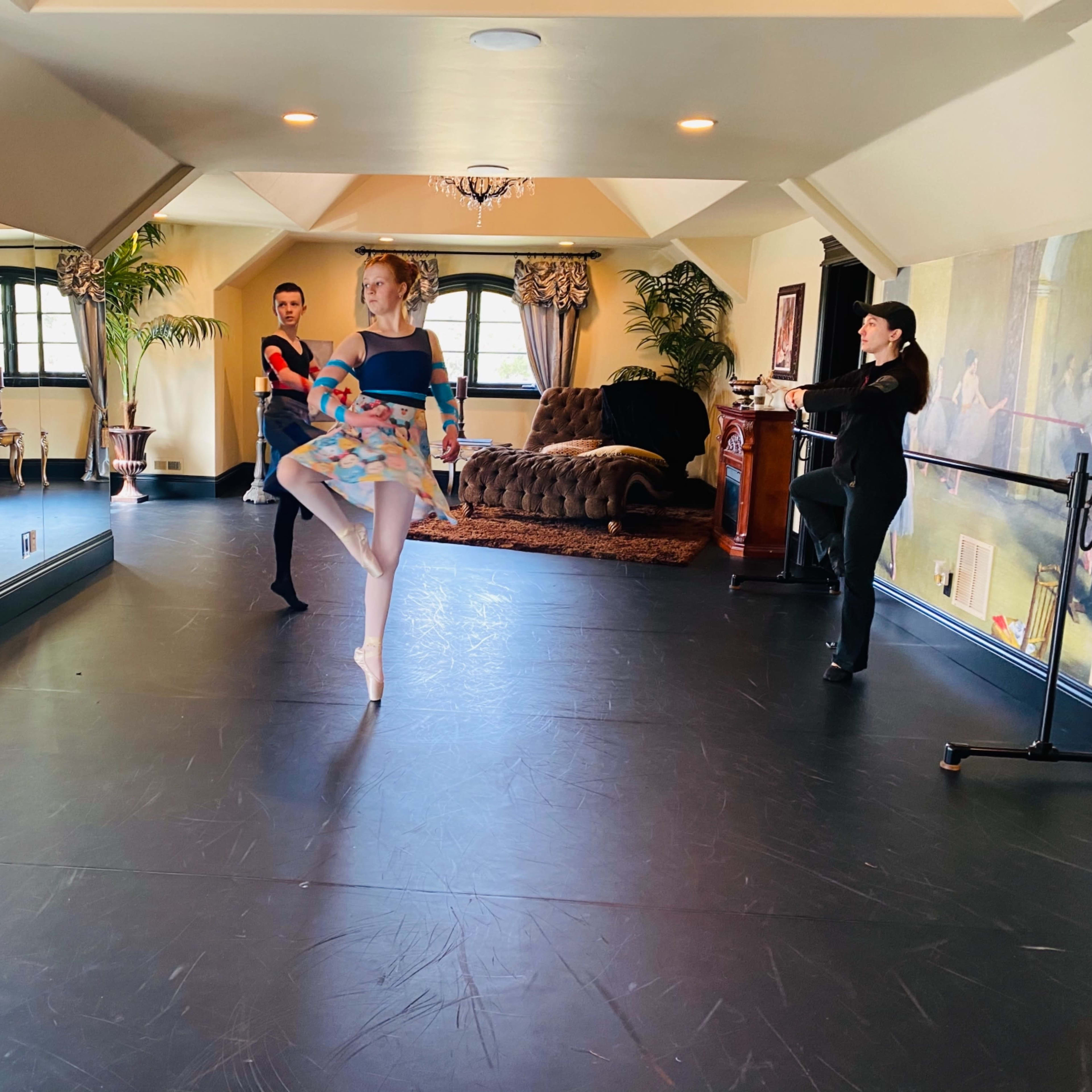A ballet dancer performs a pose in a studio while two instructors observe in the background.