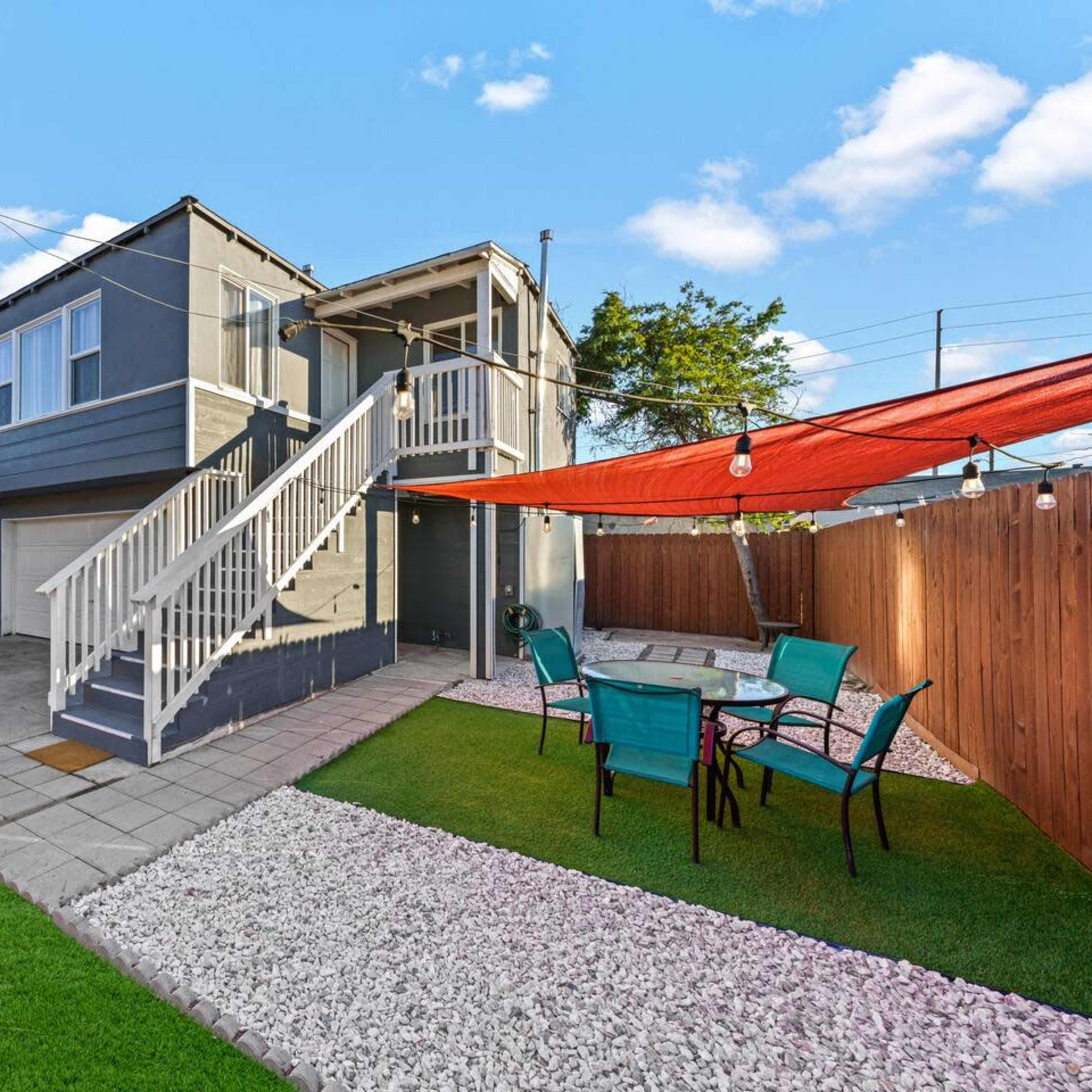 A two-story gray house with a staircase leading to the upper level, set in a yard featuring a patio area with outdoor chairs and a gravel pathway, surrounded by wooden fencing.