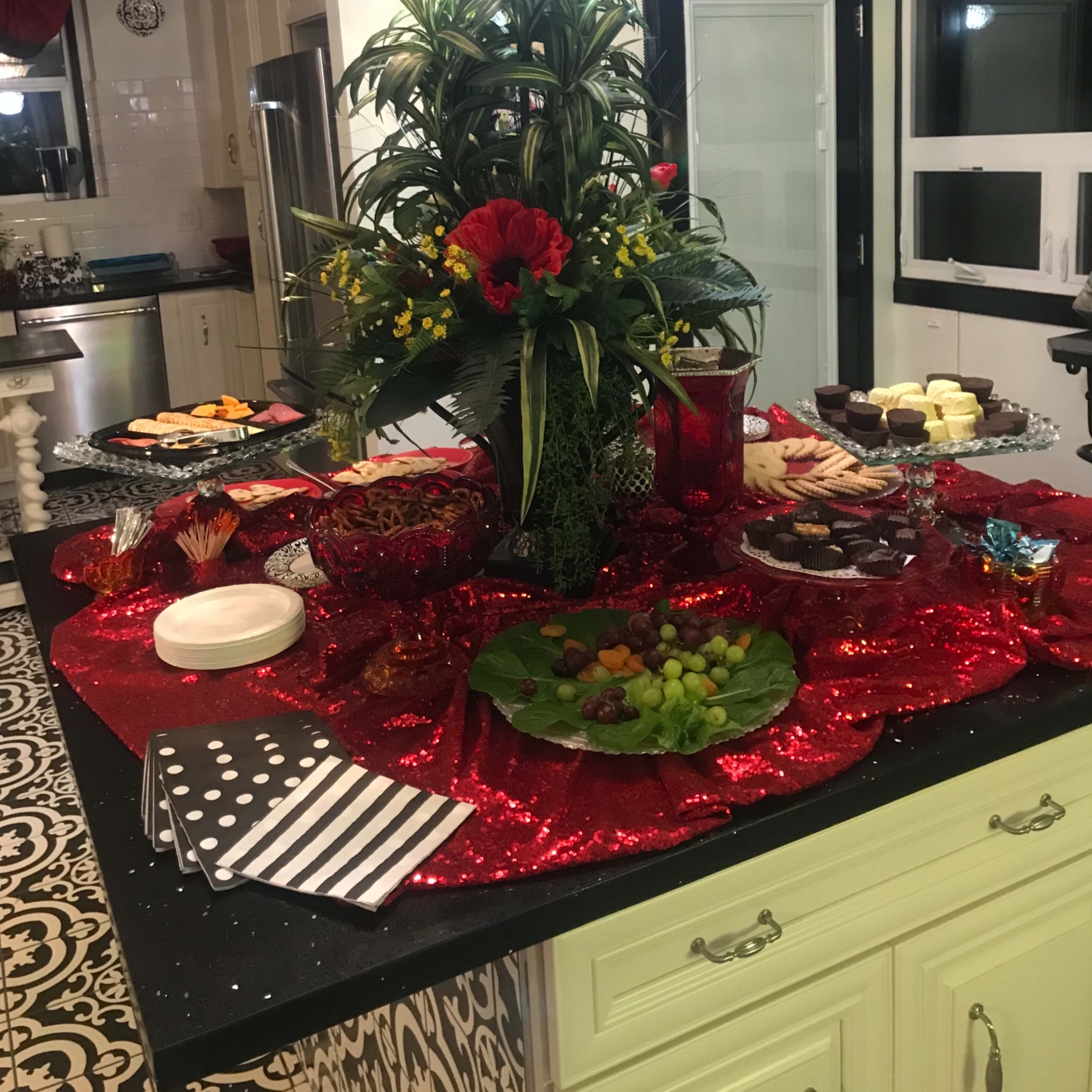 A decorative kitchen island adorned with a large floral centerpiece and various dishes of appetizers and desserts arranged on a red sparkly tablecloth.