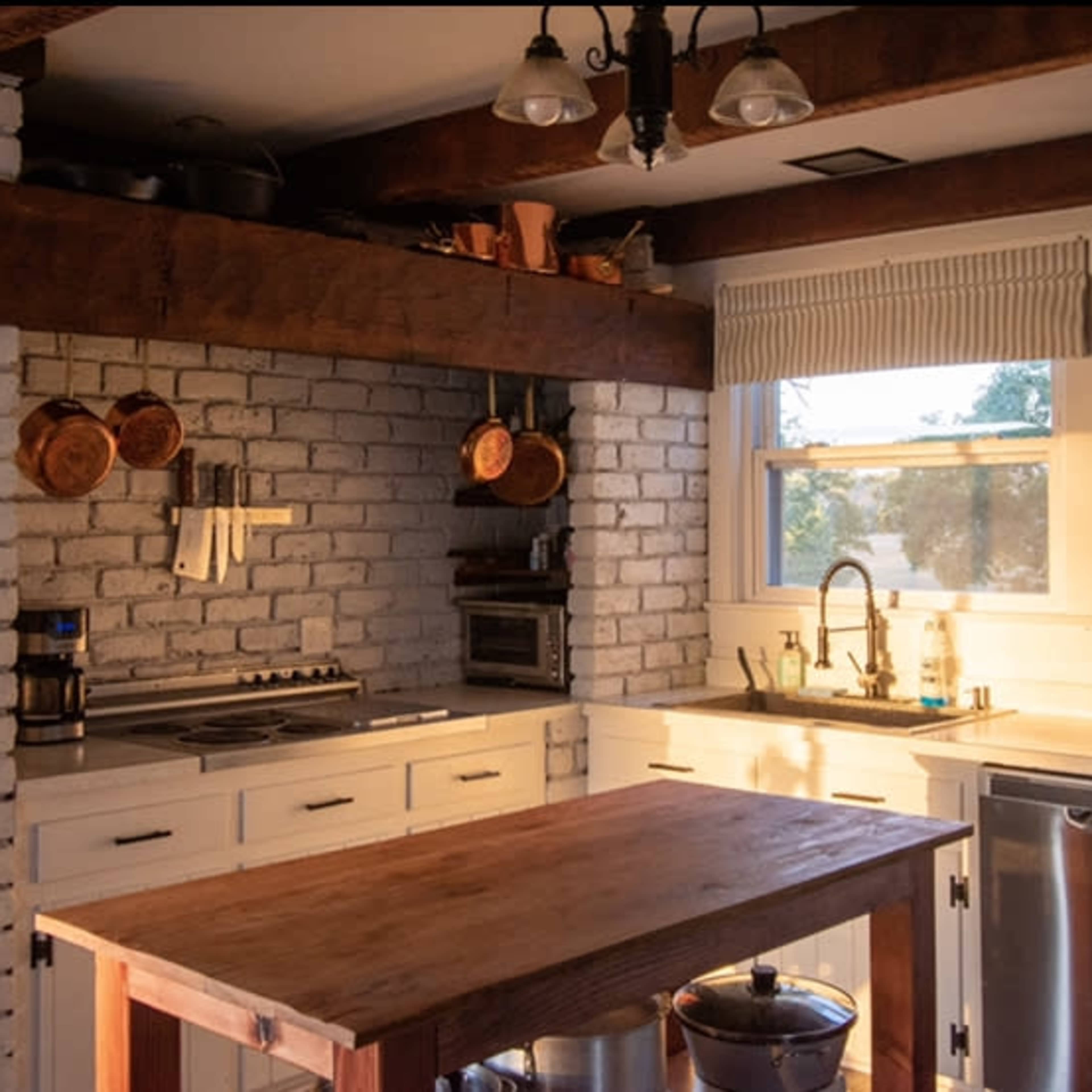 A kitchen with white brick walls, wooden beams, and a central wooden island with various kitchen appliances and utensils displayed.