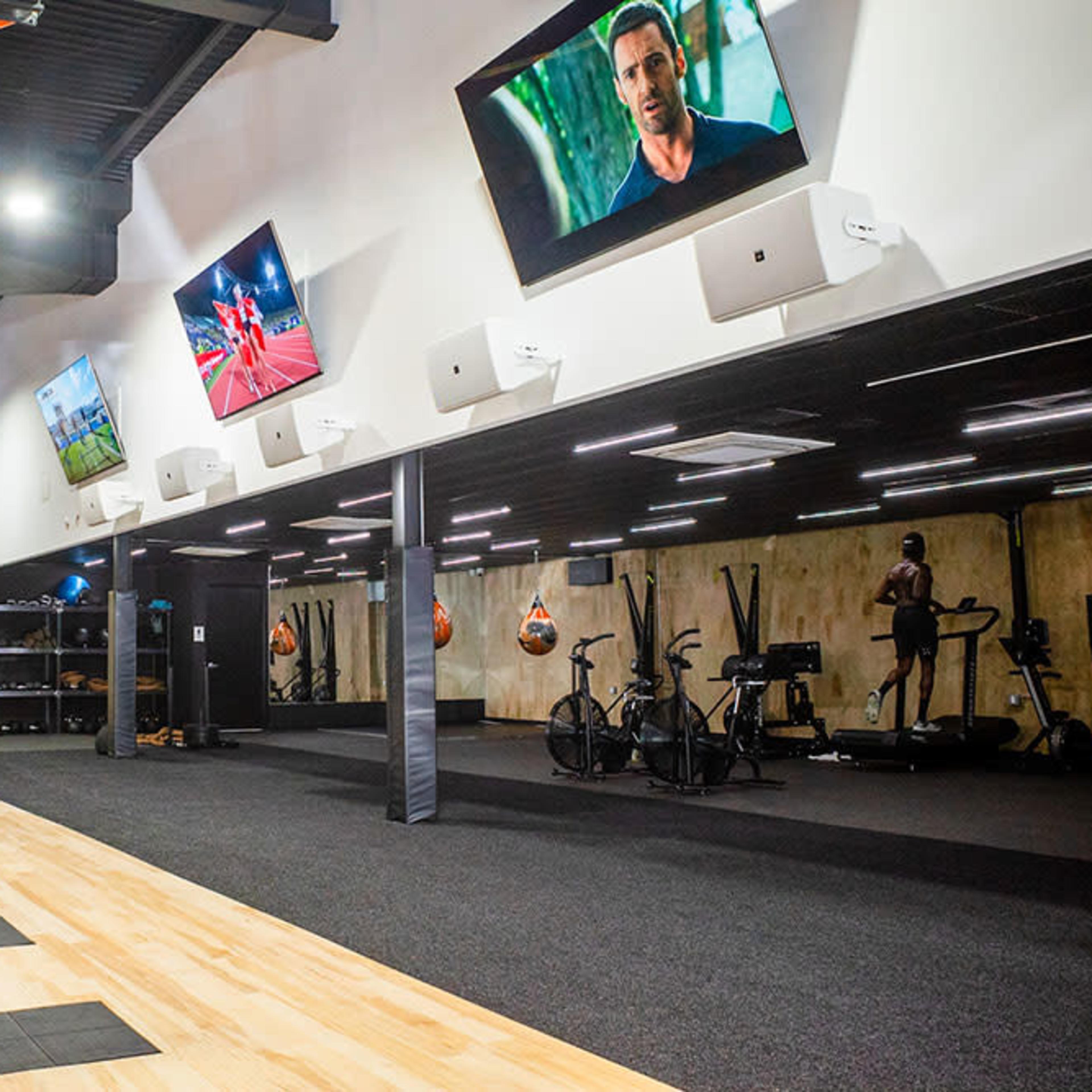 A modern fitness gym with various workout stations, including exercise bikes and a section for strength training, illuminated by overhead lights and featuring multiple screens on the walls.