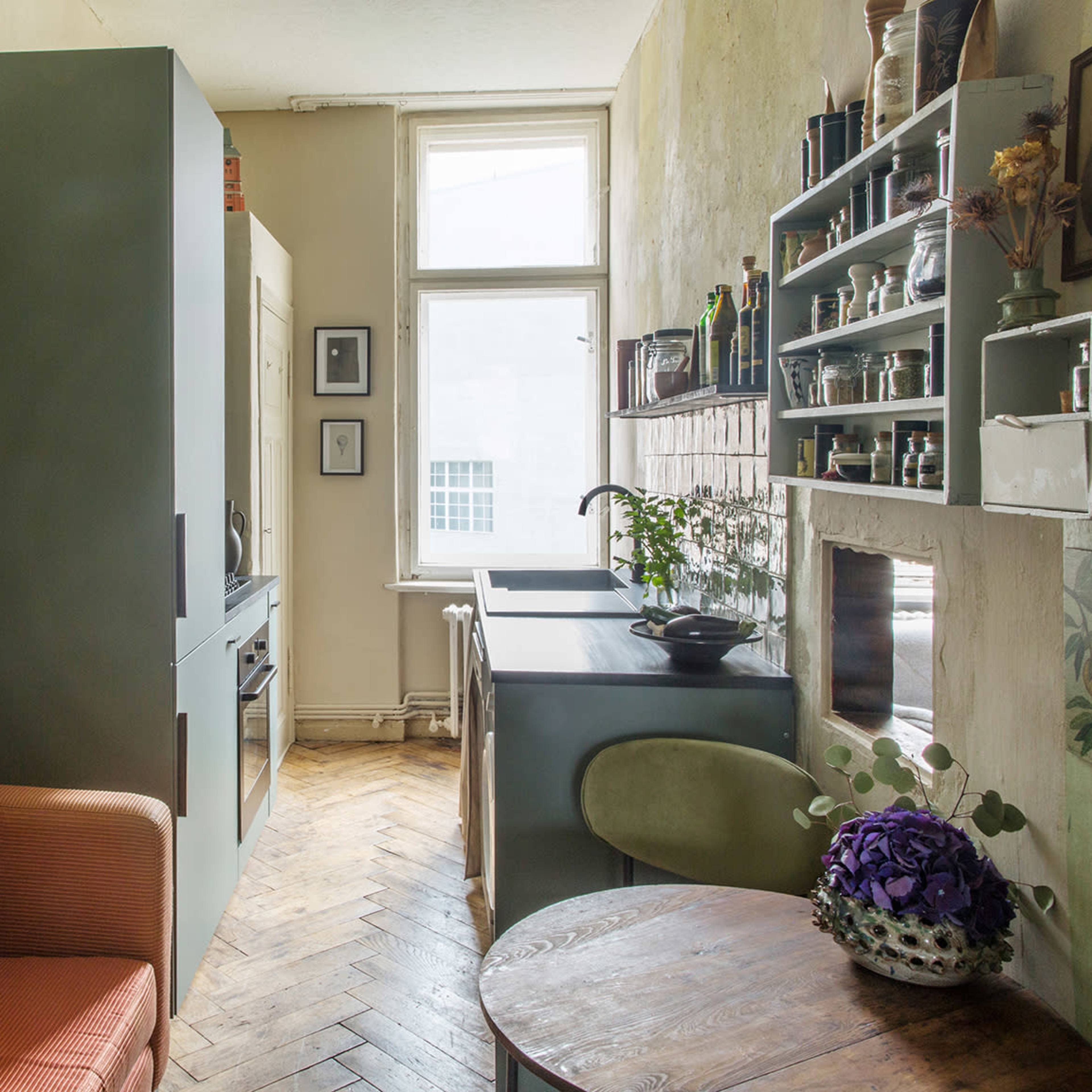 A compact kitchen and living area featuring a green kitchen unit, a small dining table, and an orange sofa near a large window.
