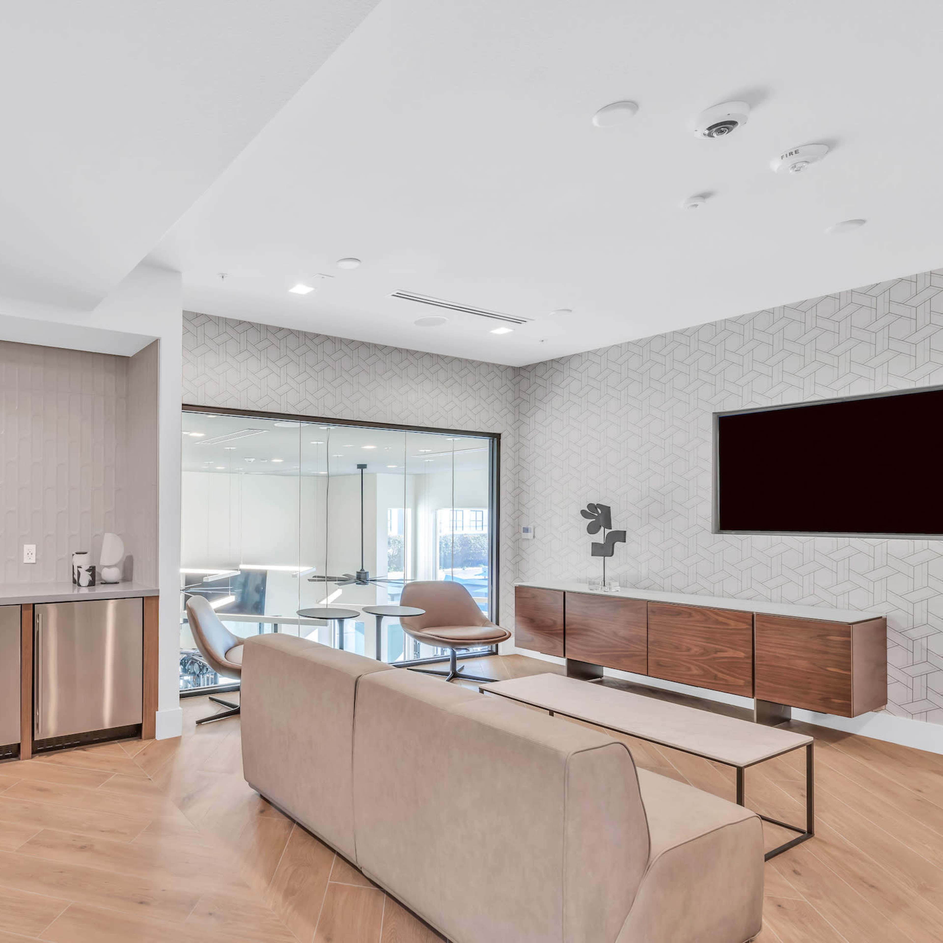 A modern lounge area featuring a kitchen space with stainless steel appliances, a beige sofa, and a glass meeting room in the background.