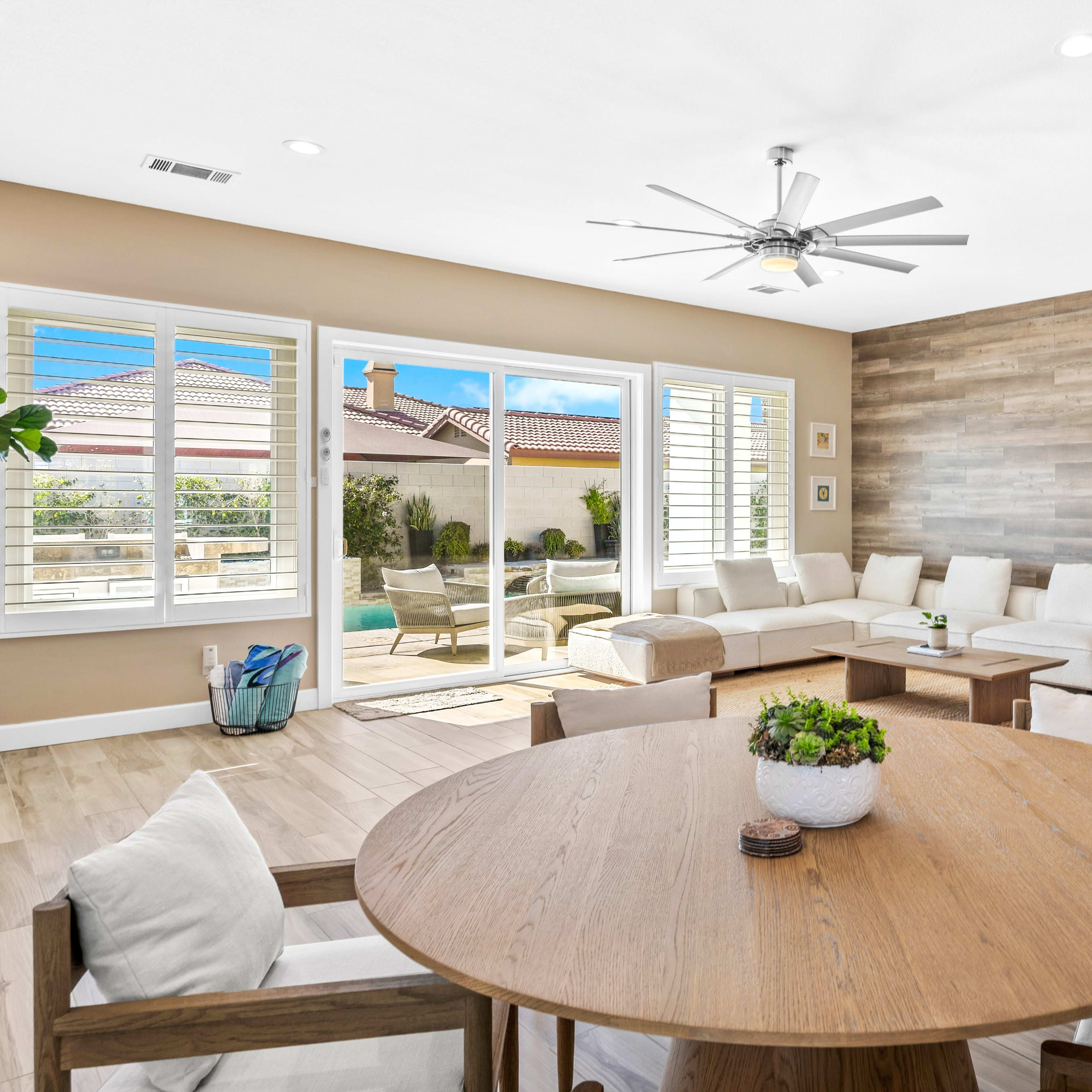 A modern living room features a circular dining table surrounded by chairs, with a sectional sofa and large windows overlooking an outdoor space.