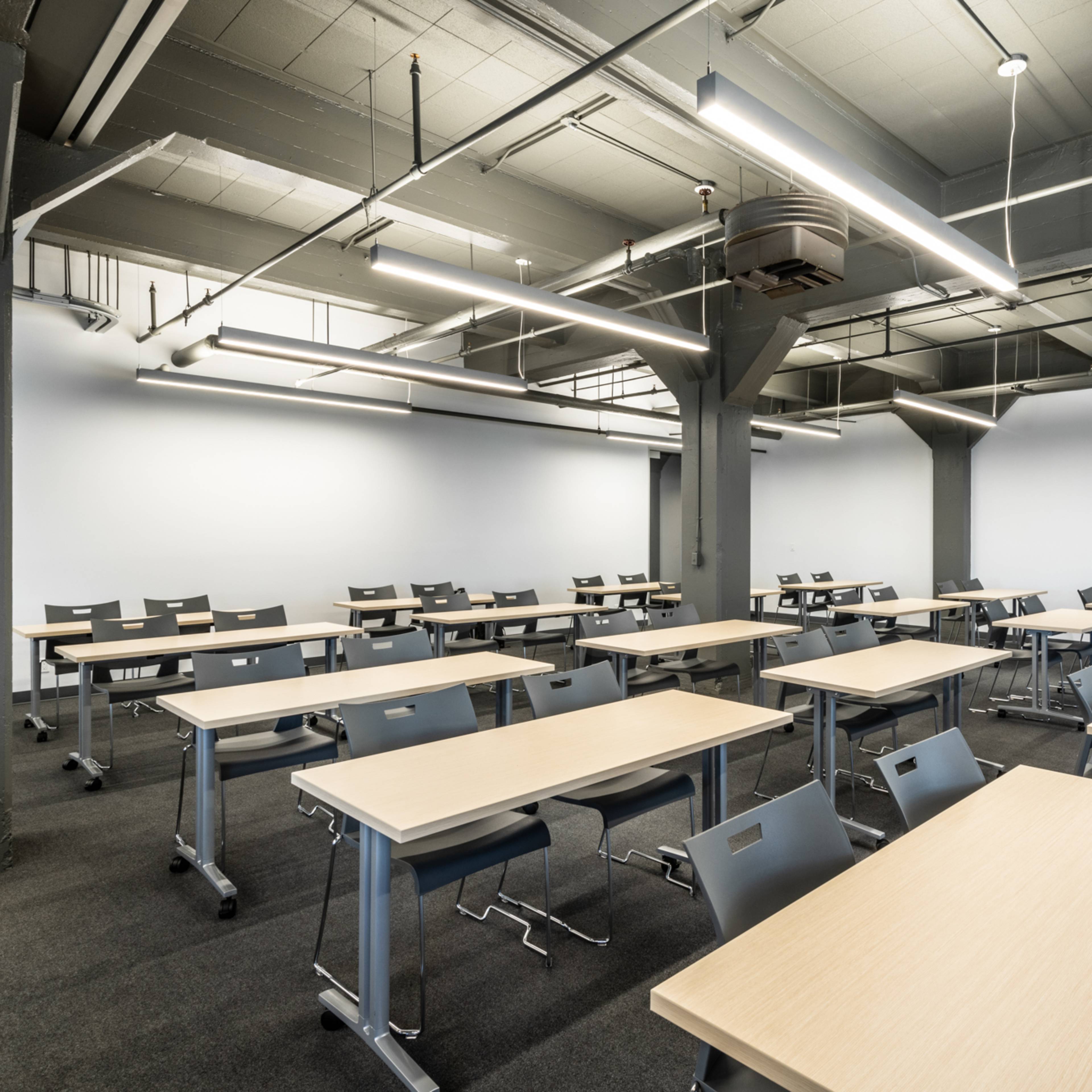 A spacious classroom featuring rows of tables and chairs arranged neatly, with gray walls and overhead lighting.