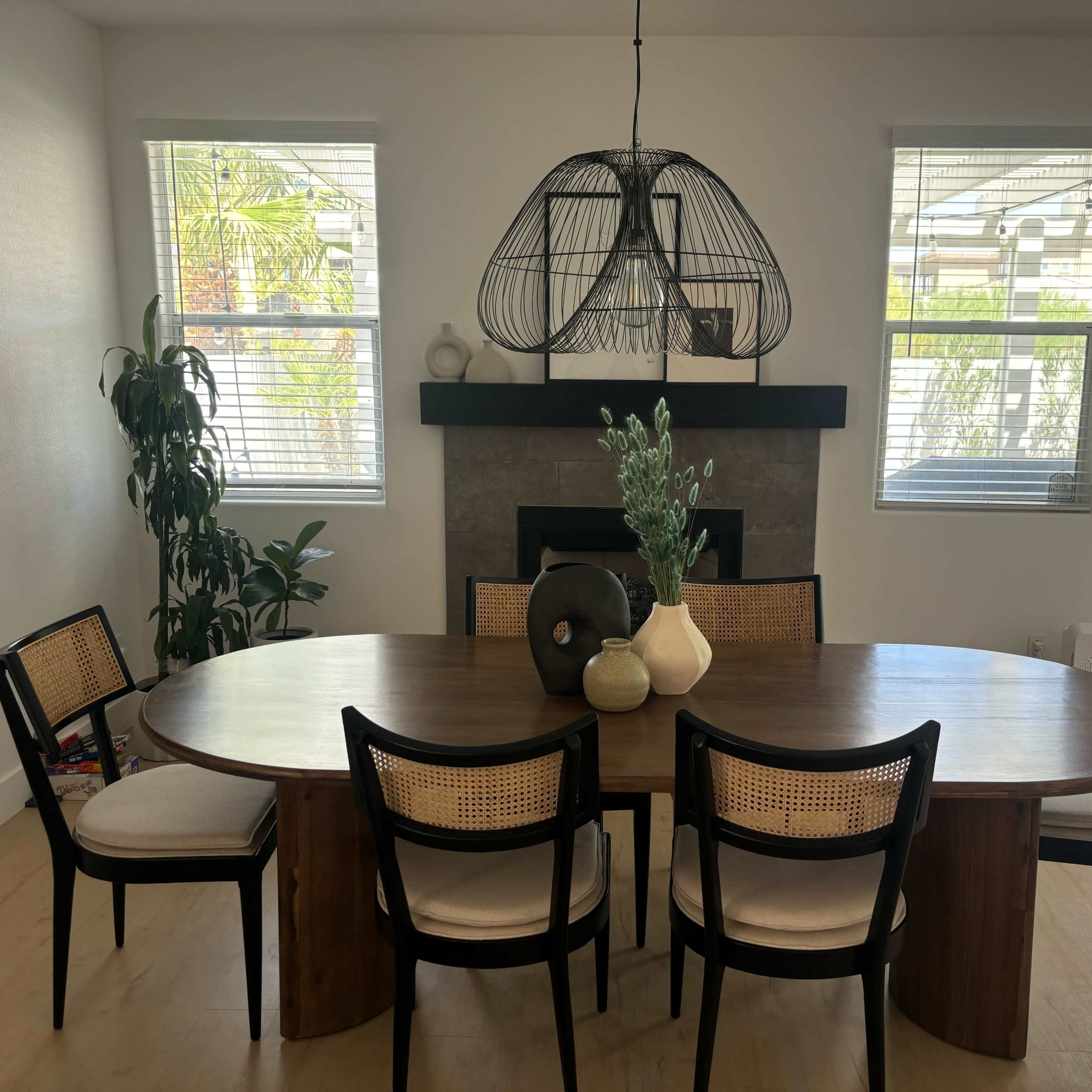 A round wooden dining table with six chairs is positioned in a bright room featuring a large pendant light and a fireplace.