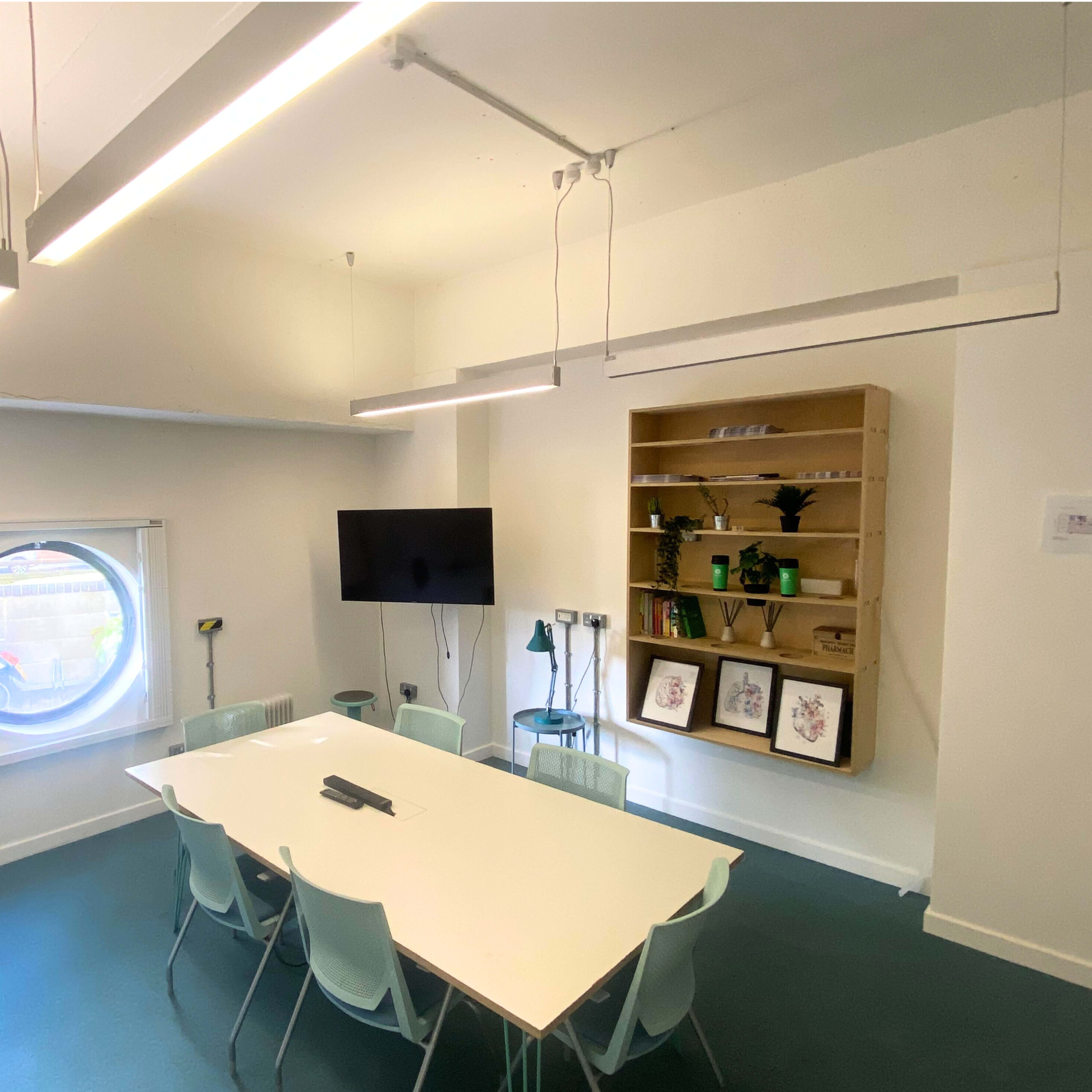 A modern meeting room featuring a large white table surrounded by chairs, a circular window, a television on the wall, and shelves with decorative items.