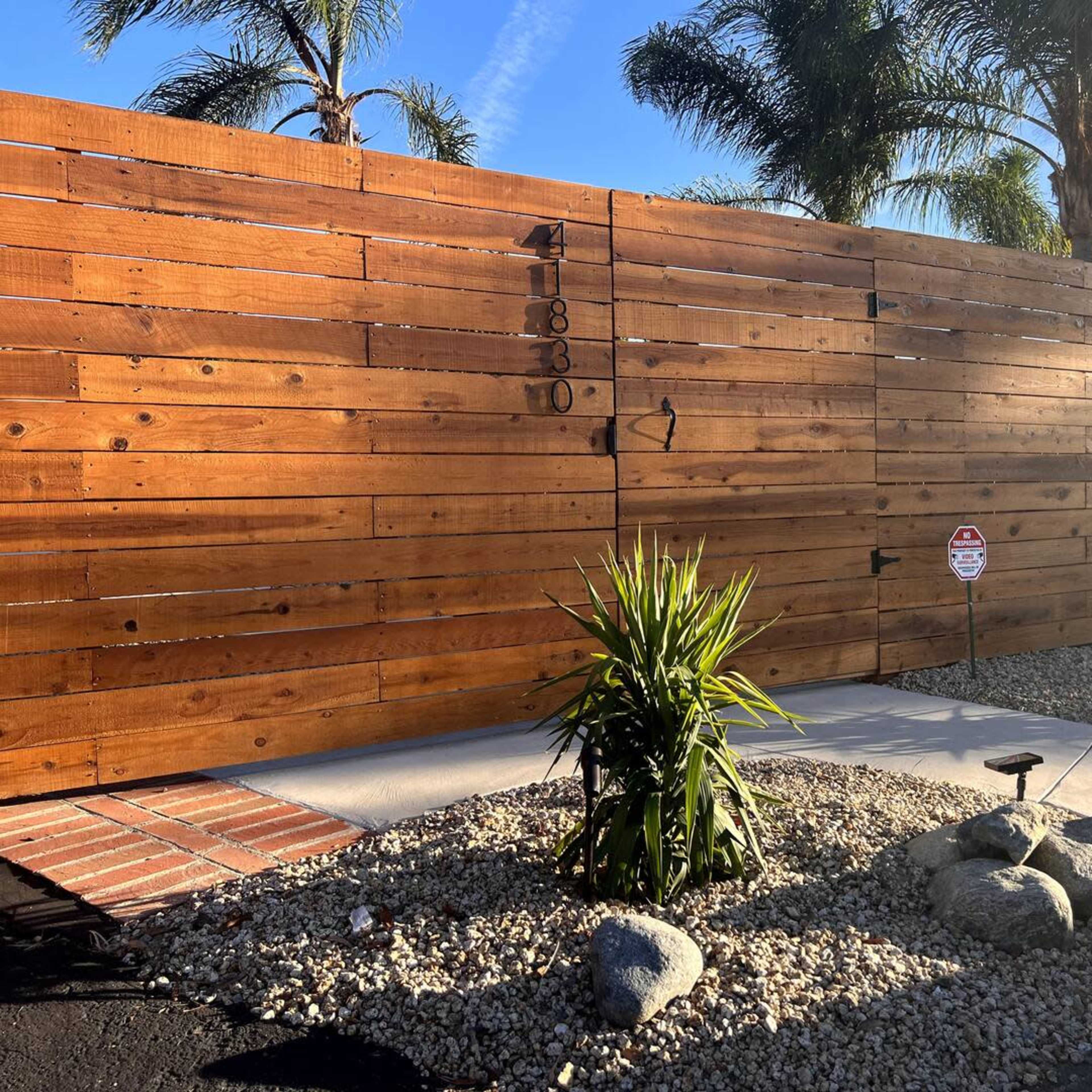 A wooden slat fence gates off a landscaped entrance with palm trees and ornamental rocks.