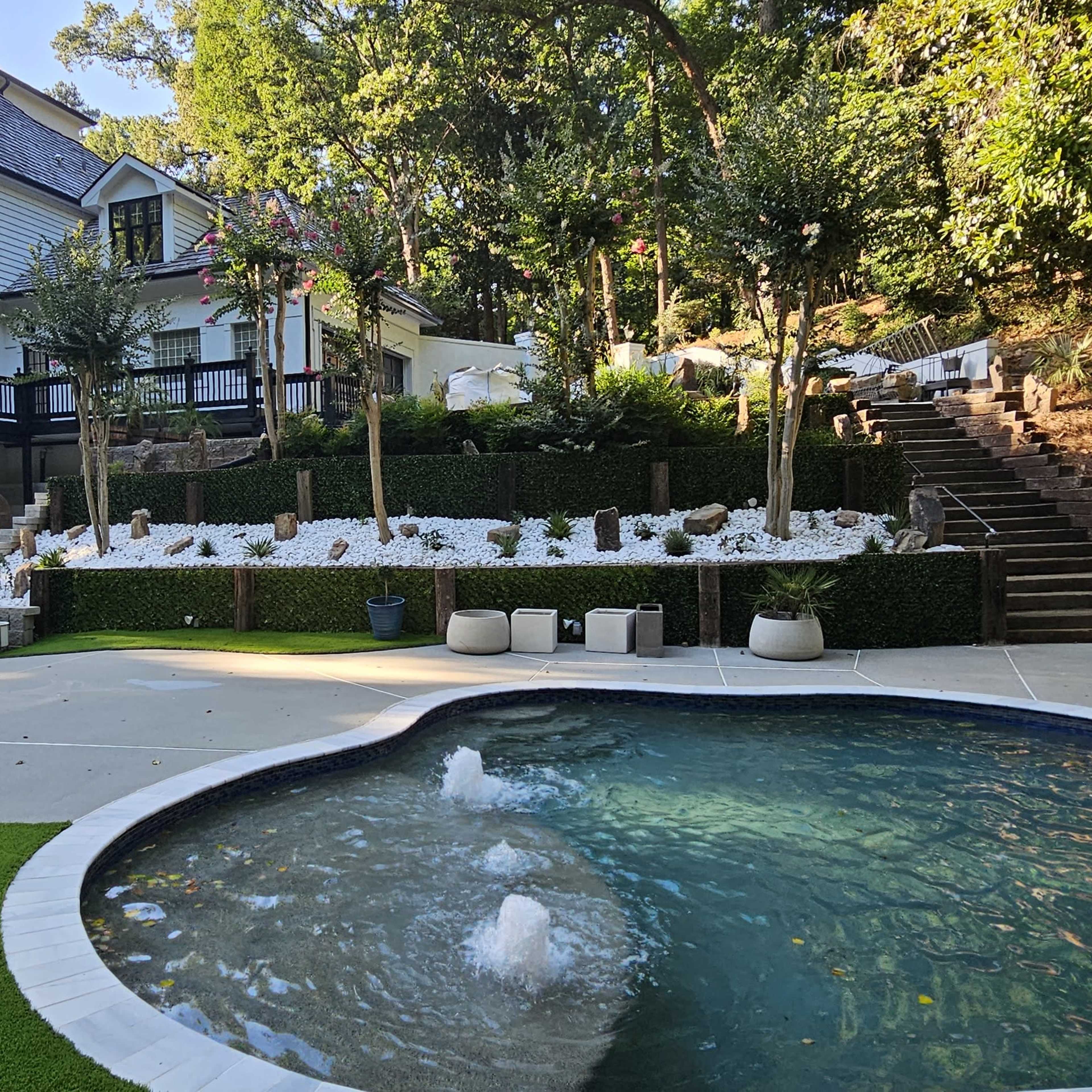 A landscaped backyard with a swimming pool, greenery, and a multi-level house surrounded by trees.