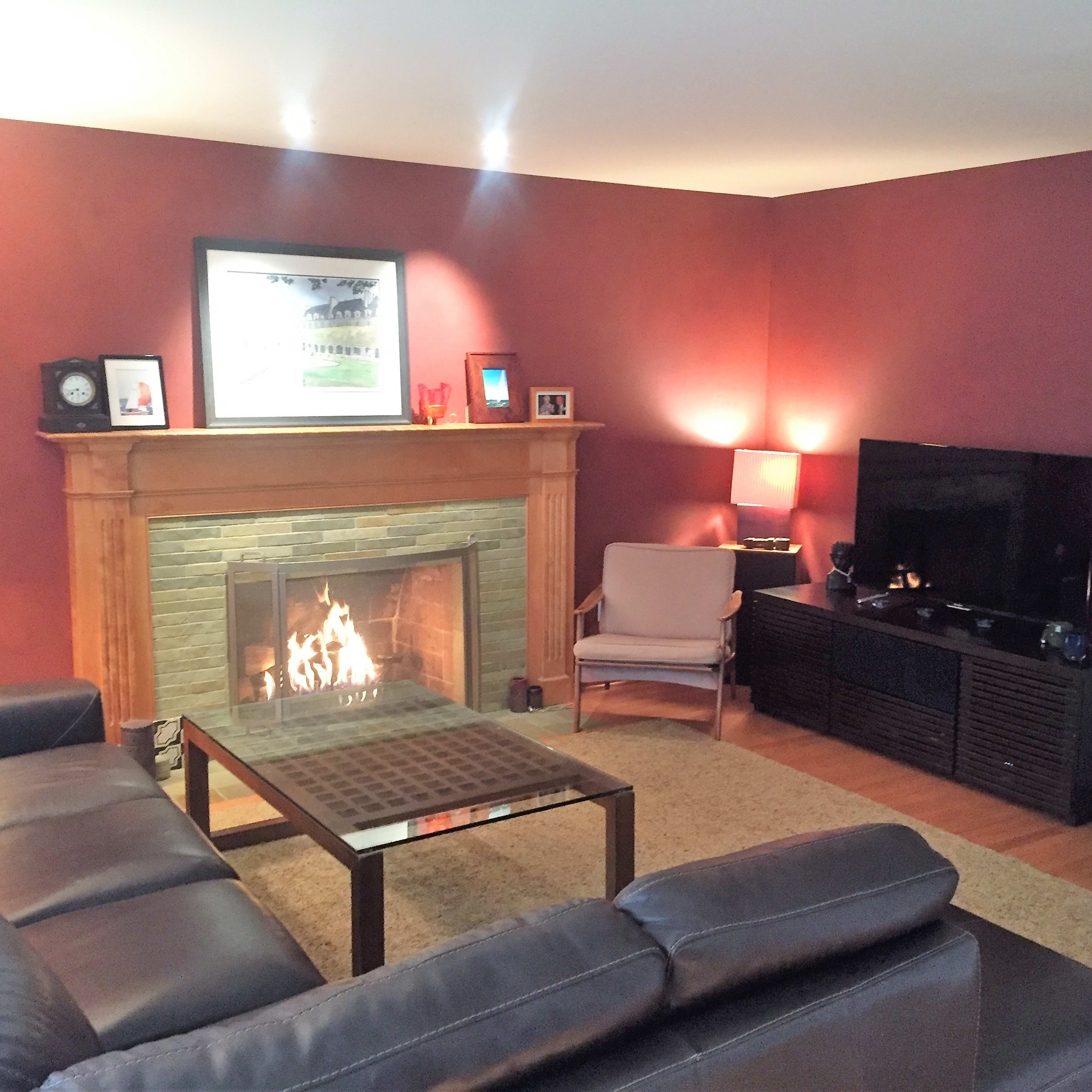 The living room features a stone fireplace with a lit fire, a large leather sectional sofa, and a television mounted on a wall across from the seating area.