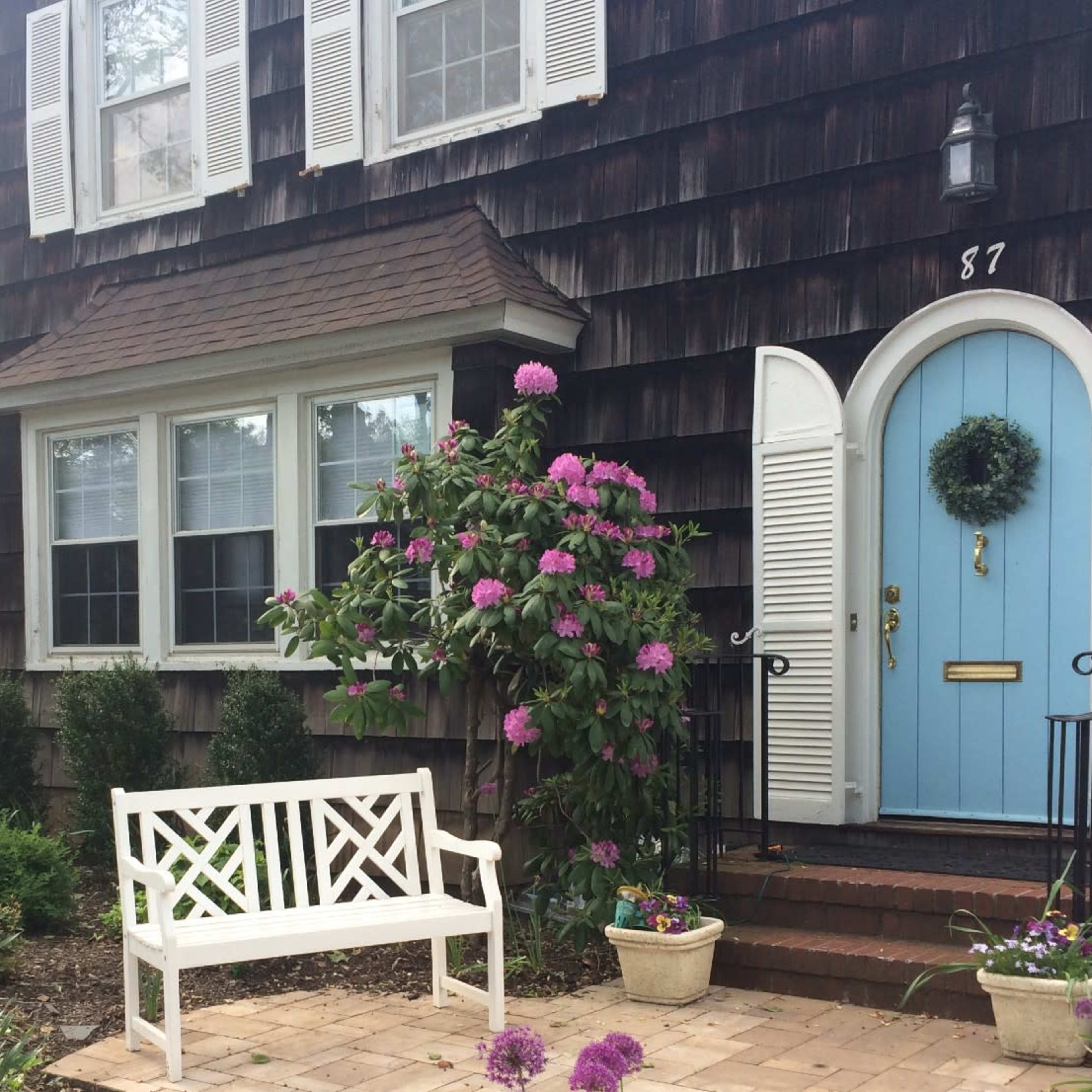 A charming house with a blue front door and flower beds filled with purple blooms in front of a wooden-shingled exterior.