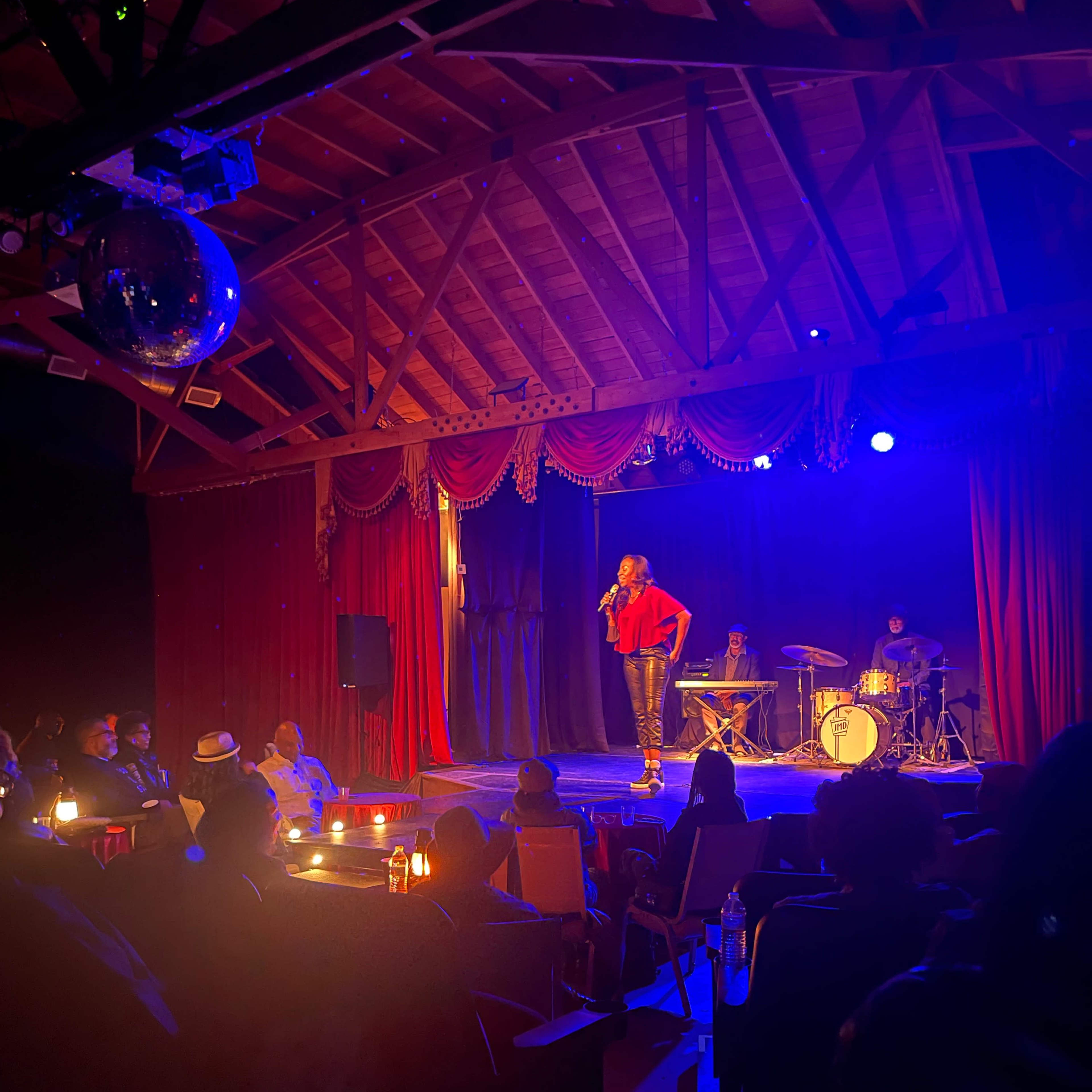 A performer stands on stage in front of an audience at a dimly lit venue with red curtains and a band setup in the background.