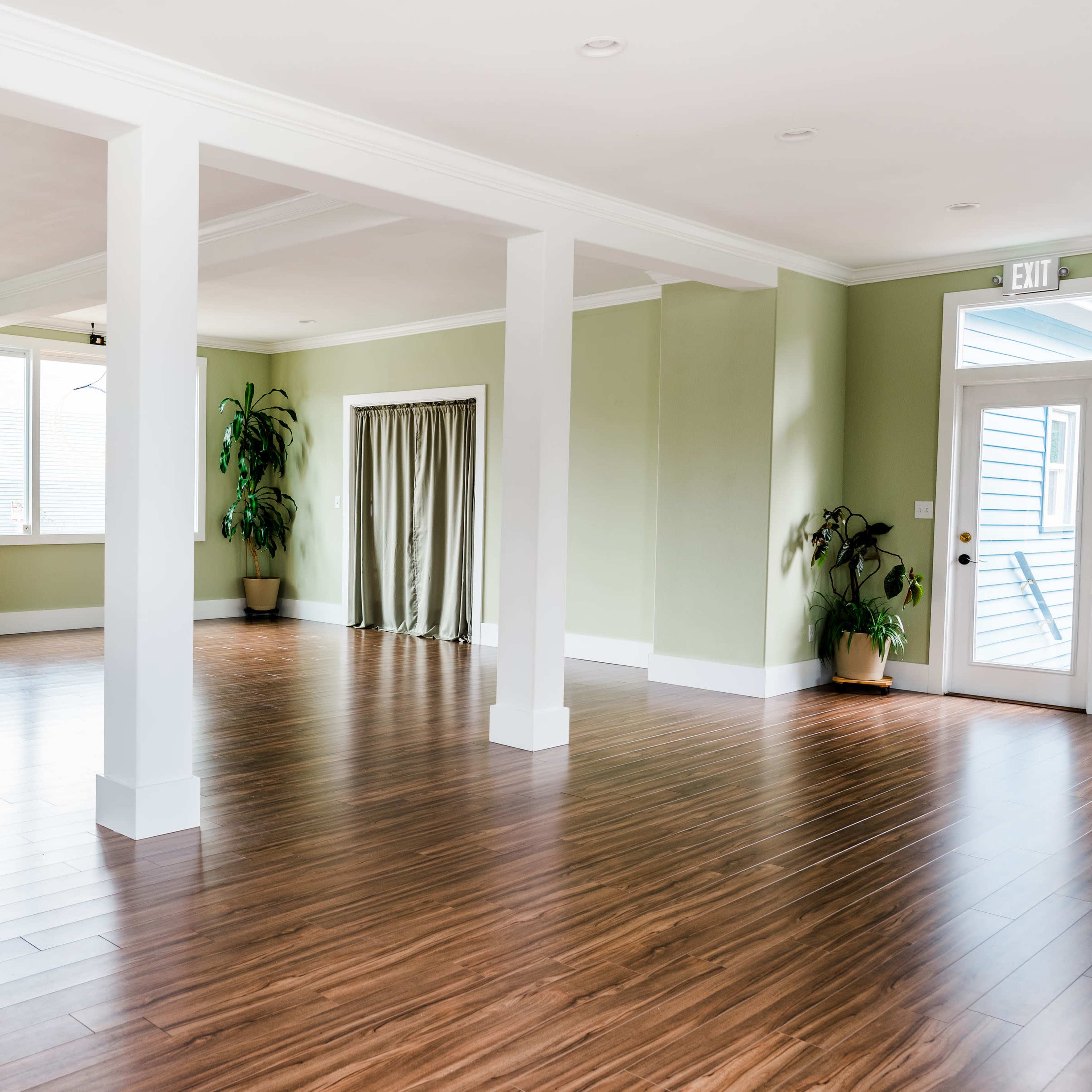 An empty room with wooden flooring, green walls, and several windows, featuring plants in the corners and a doorway leading outside.