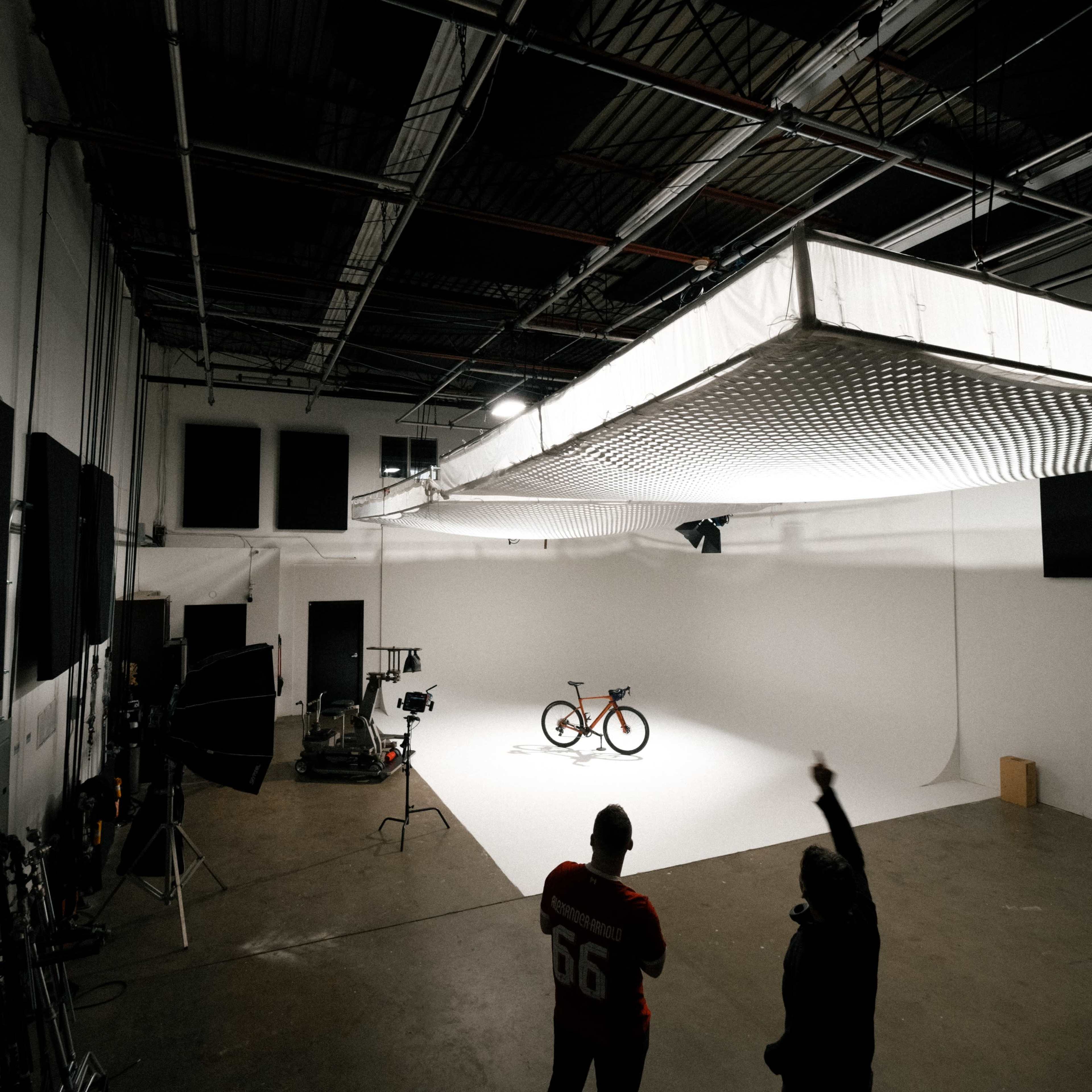 Two individuals stand in a photography studio, observing a bicycle positioned on a white backdrop under bright lights.