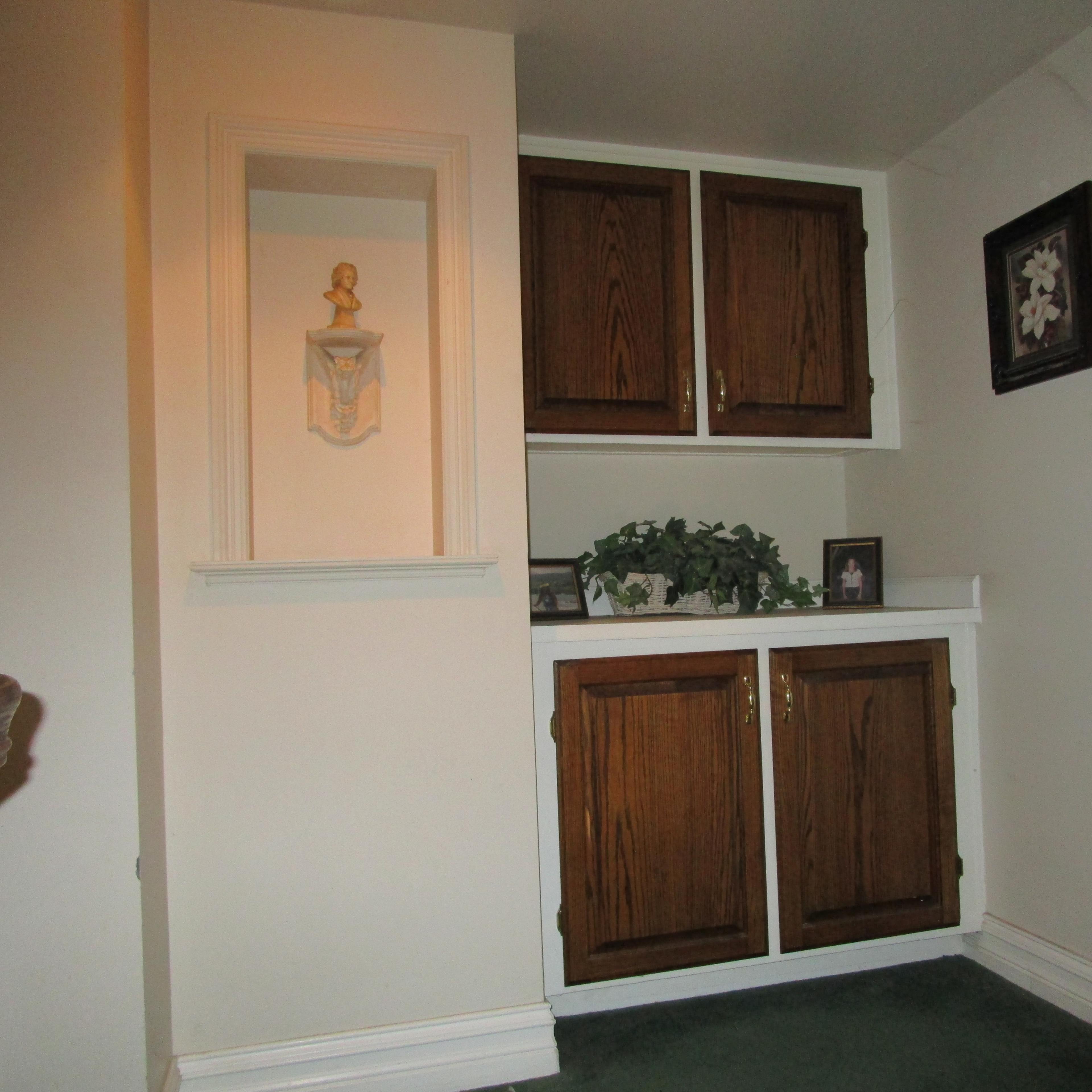 A small hallway nook with two wooden cabinet doors below a shelf that holds plants and framed pictures, all against a wall painted in neutral tones.