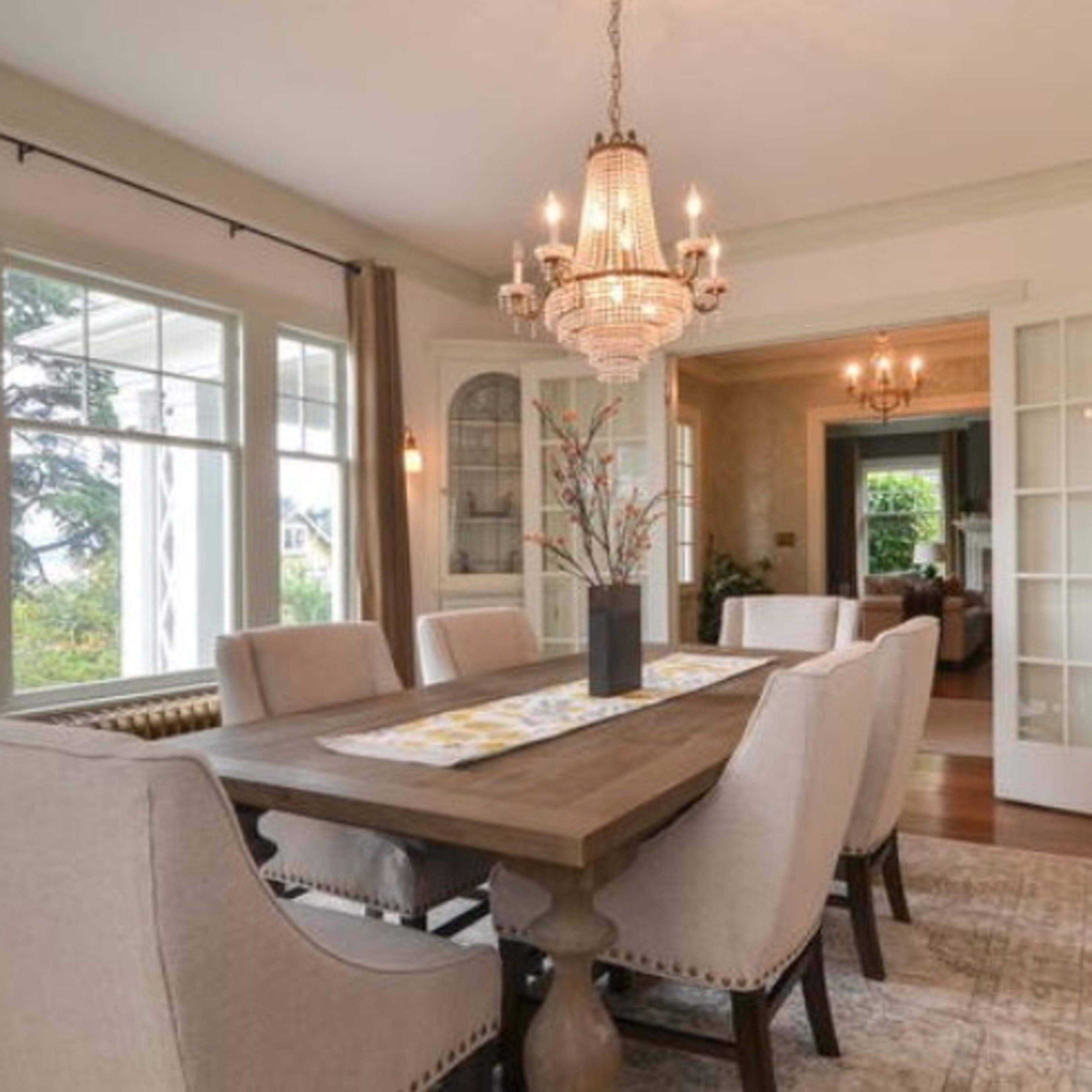 A dining room features a wooden table surrounded by upholstered chairs, with large windows allowing natural light and a chandelier above.
