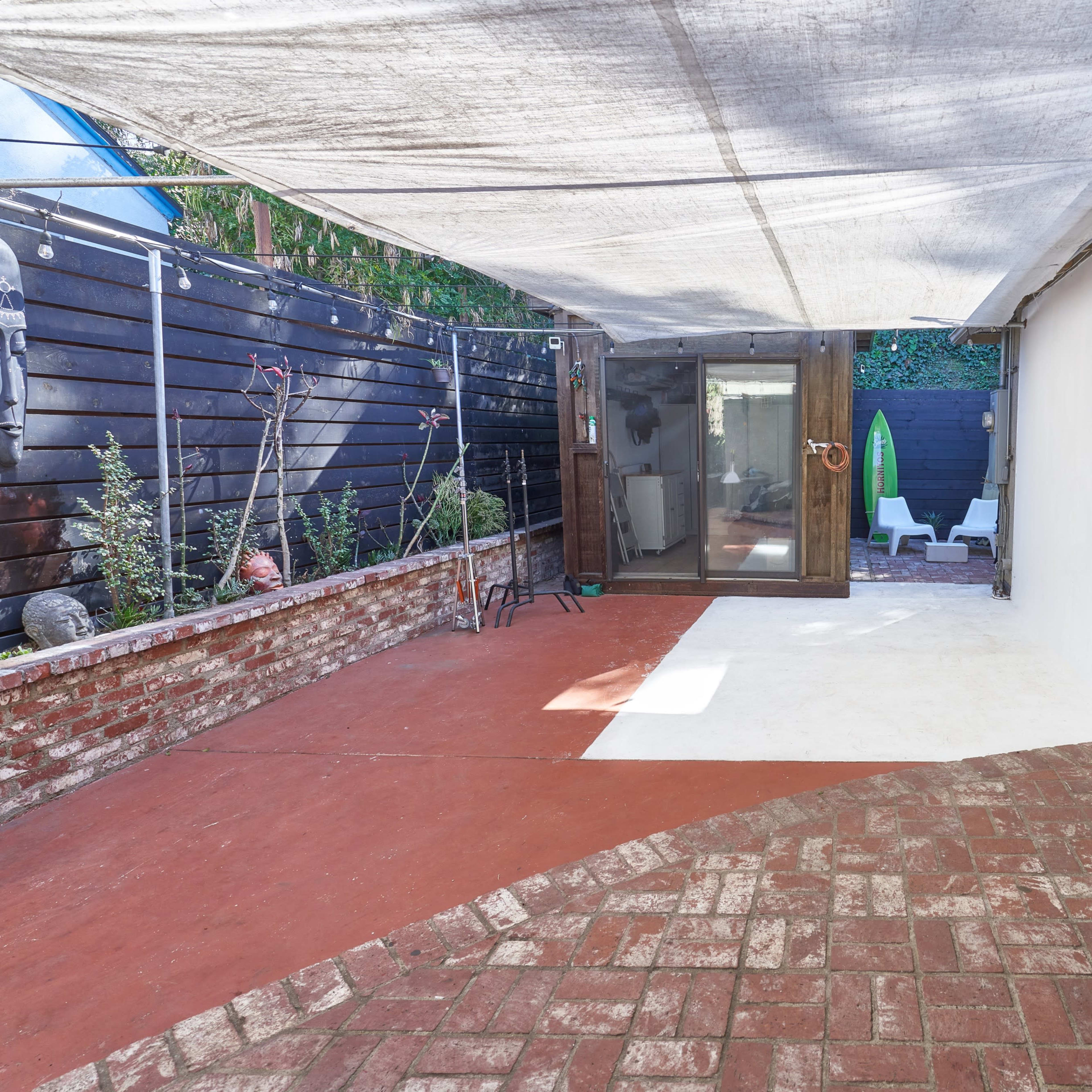 A covered outdoor space with a brick floor, featuring a glass door leading to a small enclosed area with seating and greenery.